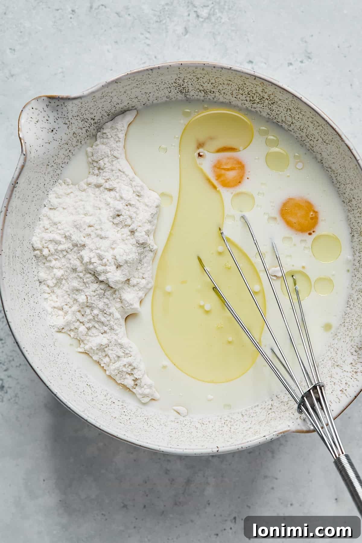 A large mixing bowl containing gluten-free pancake mix, almond milk, eggs, and avocado oil, being vigorously whisked to combine all ingredients into a smooth batter.