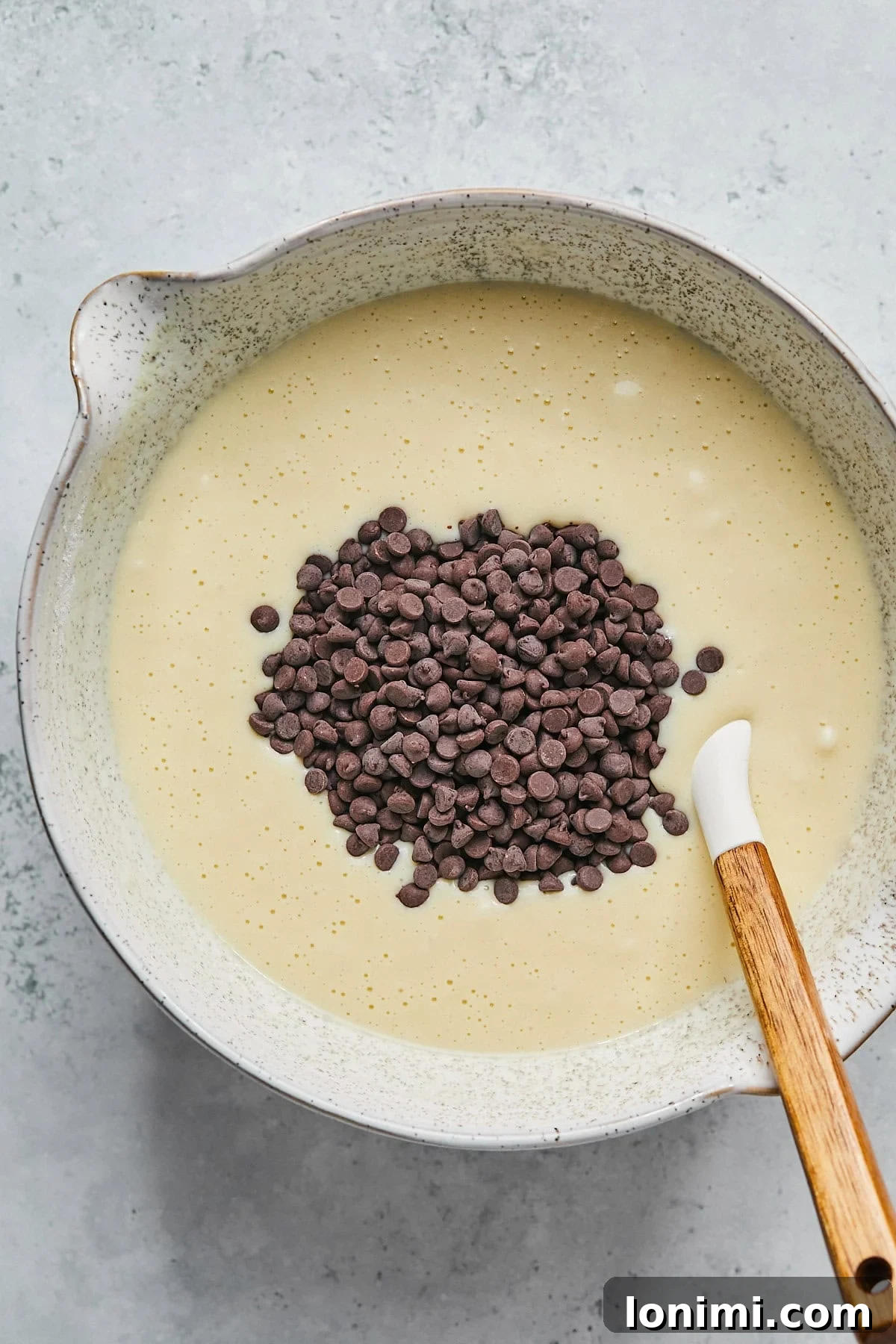 A spatula gently folding mini chocolate chips into the freshly prepared muffin batter in a white mixing bowl, ensuring even distribution.