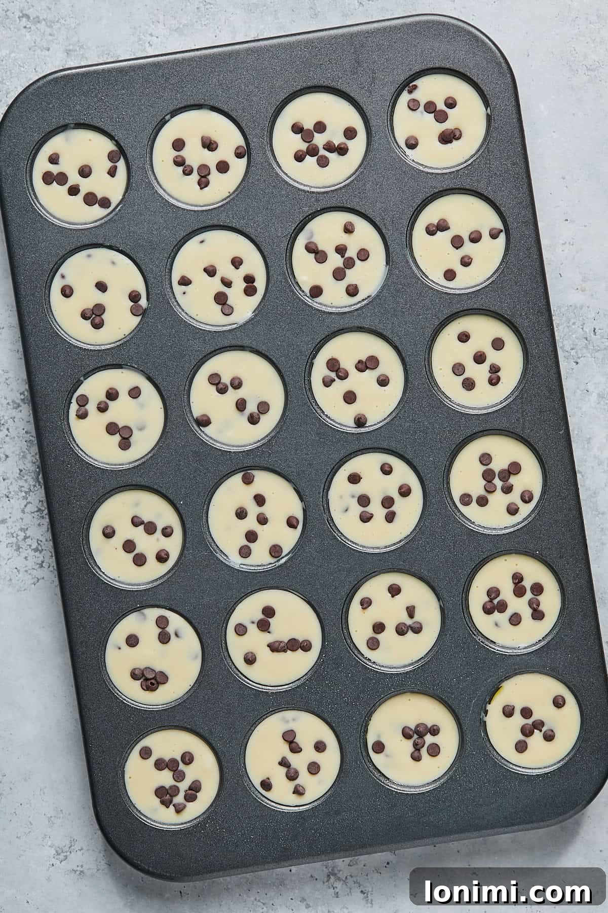 A 24-cup mini muffin pan with each cup filled almost to the brim with muffin batter and topped with a scattering of extra mini chocolate chips before baking.