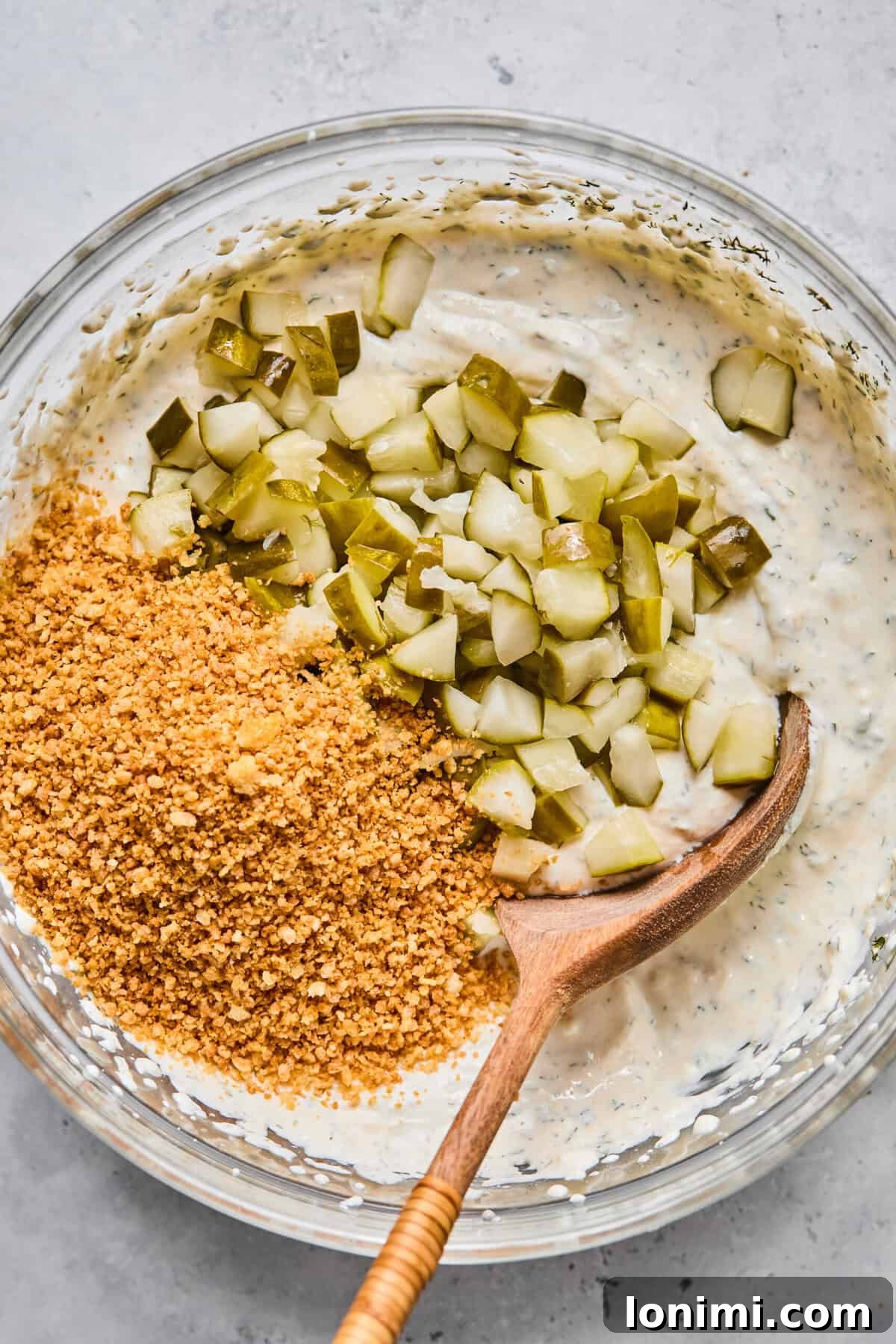 A close-up shot of chopped dill pickles and golden toasted panko breadcrumbs being gently folded into a creamy dip mixture in a bowl.