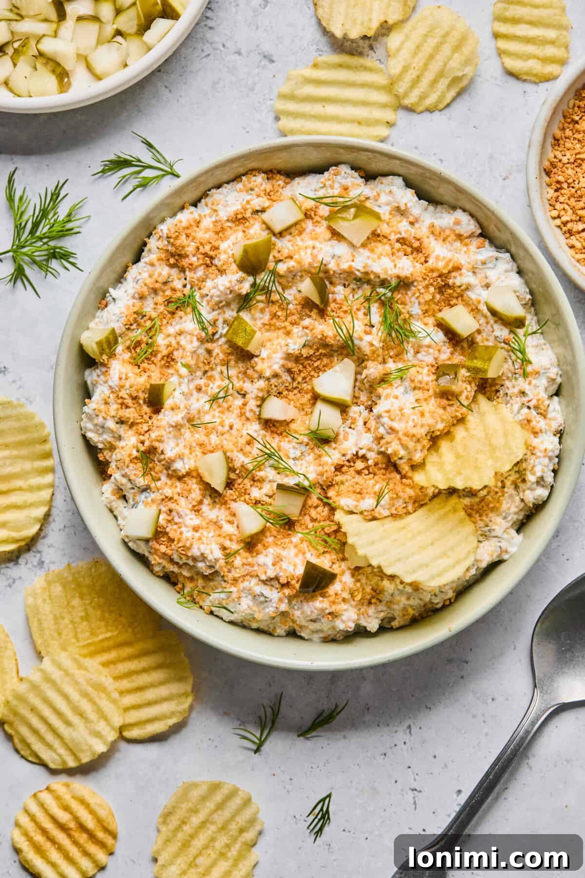 A generous serving of creamy fried pickle dip, beautifully topped with golden gluten-free panko breadcrumbs and fresh dill, surrounded by an assortment of crispy potato chips in a vibrant setting.