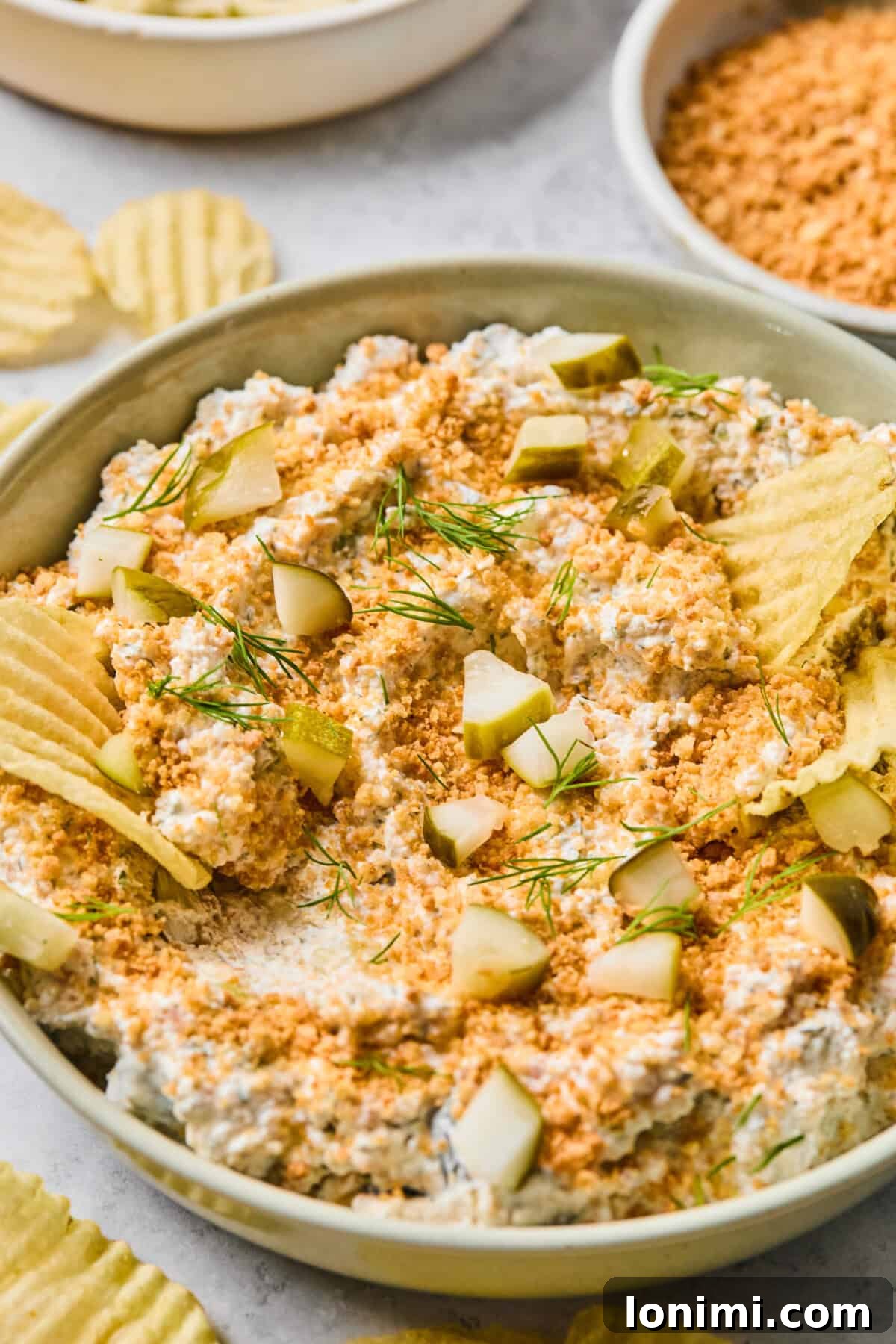 A delightful side view of a bowl brimming with creamy fried pickle dip, garnished with fresh dill and crunchy panko, accompanied by an assortment of crispy potato chips.