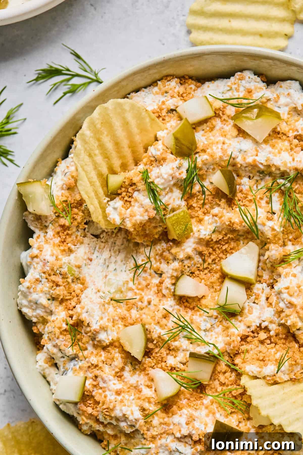 A mouthwatering close-up of creamy fried pickle dip, garnished with fresh dill and crunchy panko, with a ripple potato chip perfectly dipped in it, ready for a bite.