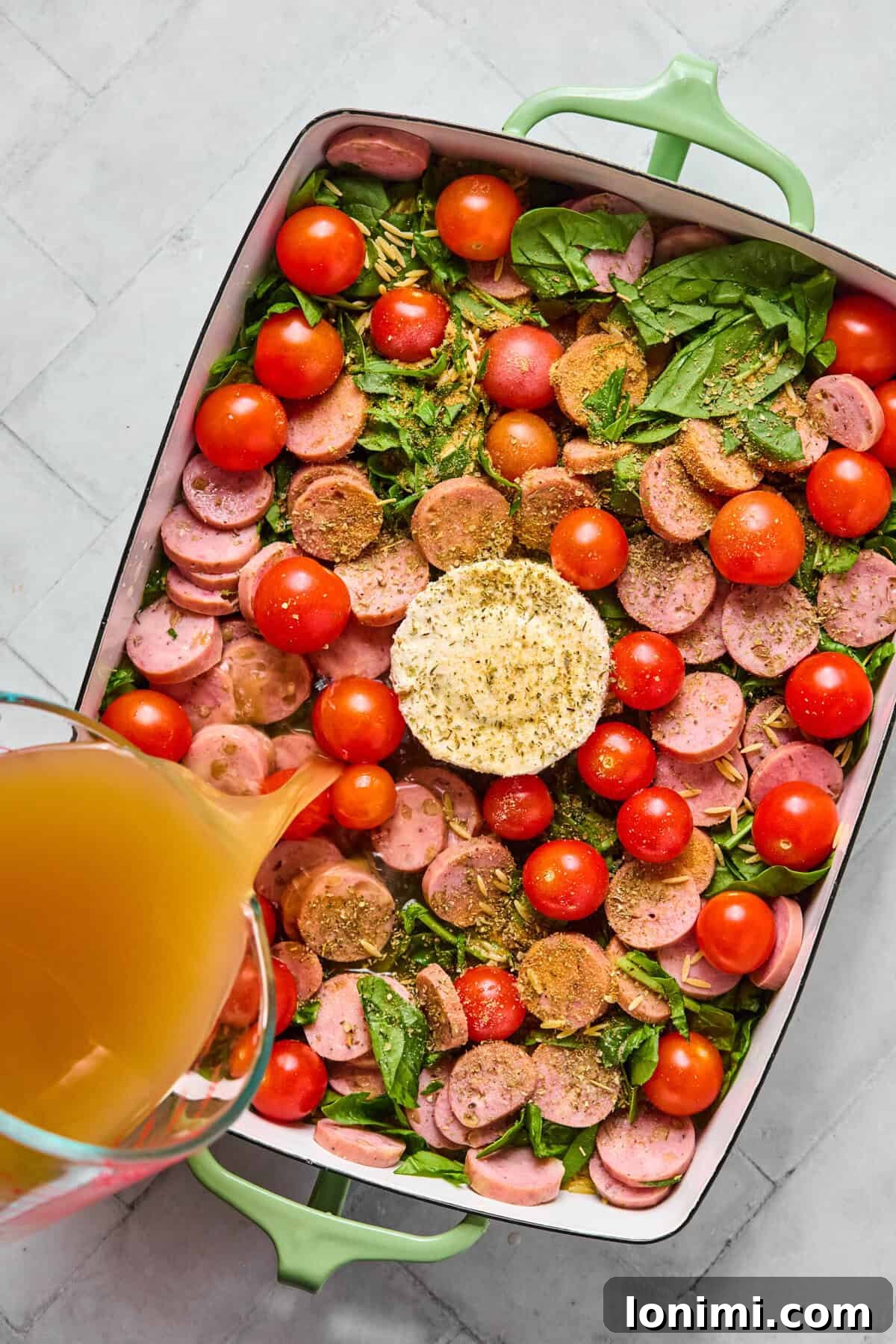 Unbaked orzo, sausage, tomatoes, and spinach arranged around Boursin cheese in a pan with broth being poured in.
