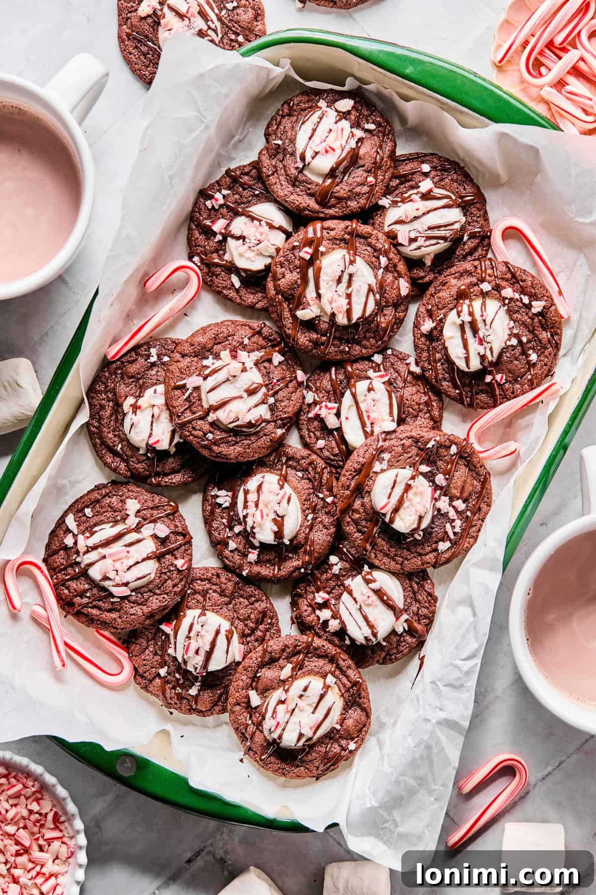 A festive arrangement of gluten-free hot cocoa cookies on a green parchment-lined tray, surrounded by mini candy canes, with a steaming mug of hot chocolate nearby.