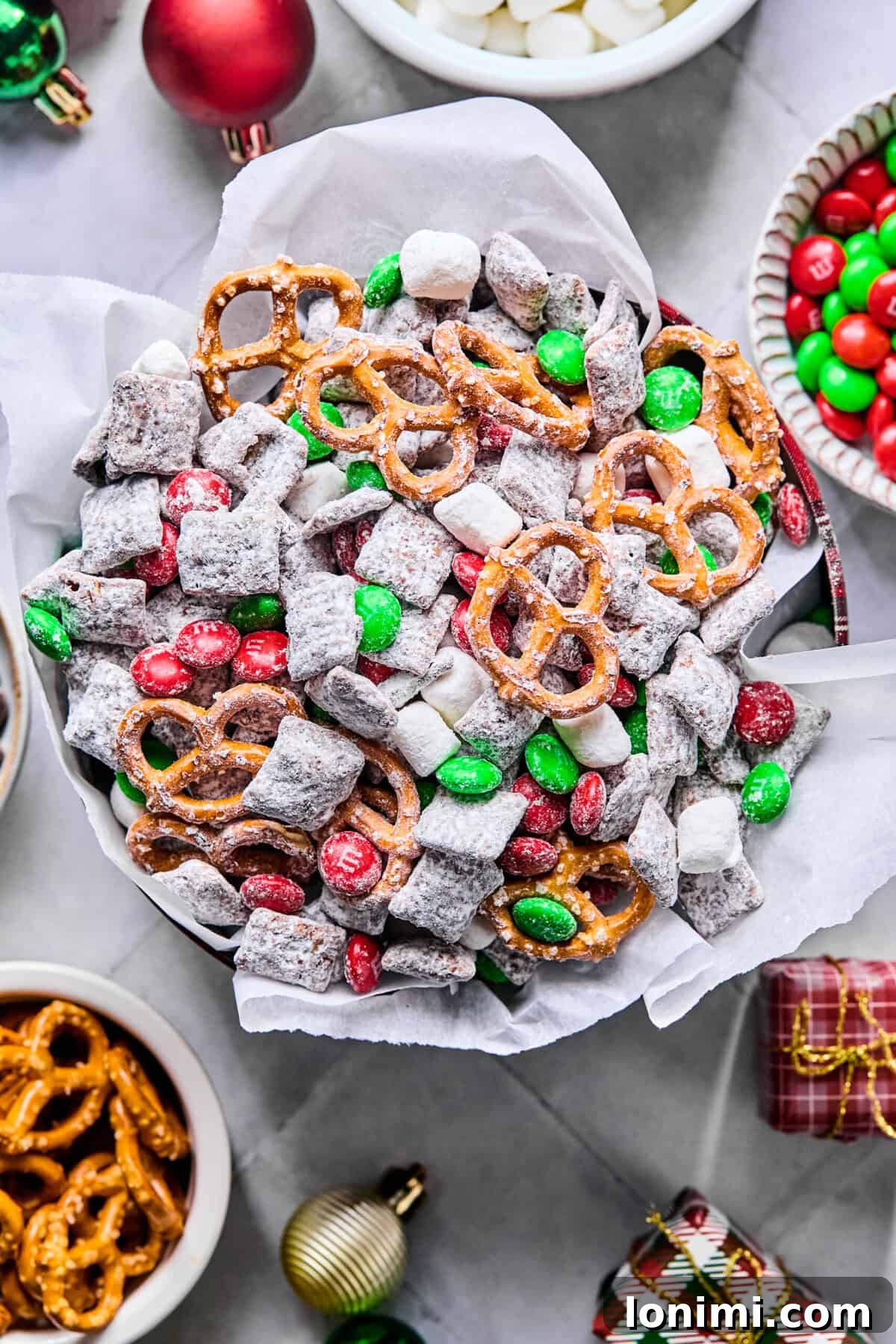 Overhead view of a festive parchment-lined serving tin filled with Christmas puppy chow on a white countertop, adorned with red and green M&Ms and mini marshmallows.