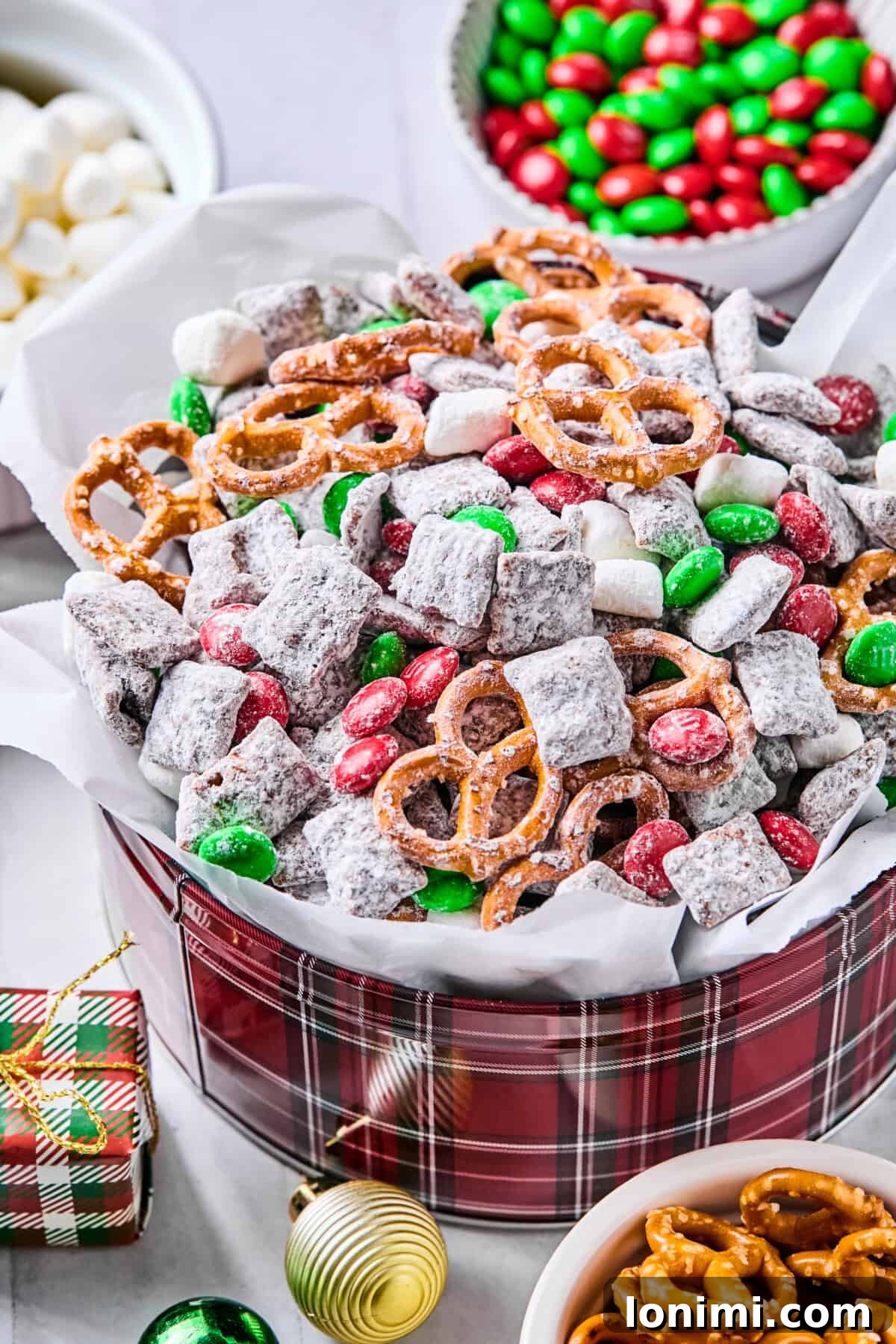 A festive parchment-lined tin filled with Christmas puppy chow, adorned with red and green M&Ms and mini marshmallows, resting on a white countertop with more candies scattered around.
