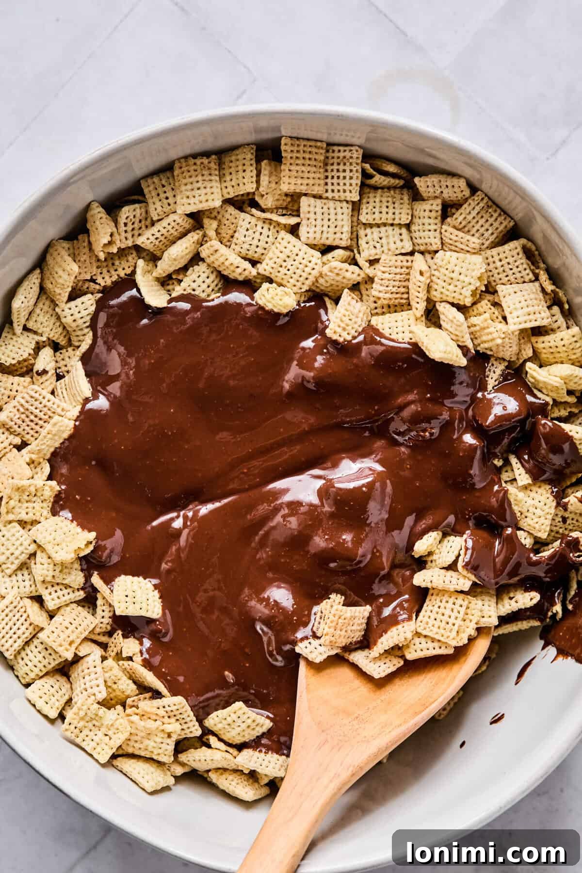 A large mixing bowl filled with crisp Rice Chex cereal, topped with the freshly melted chocolate and peanut butter mixture, with a wooden spoon resting inside ready to mix.