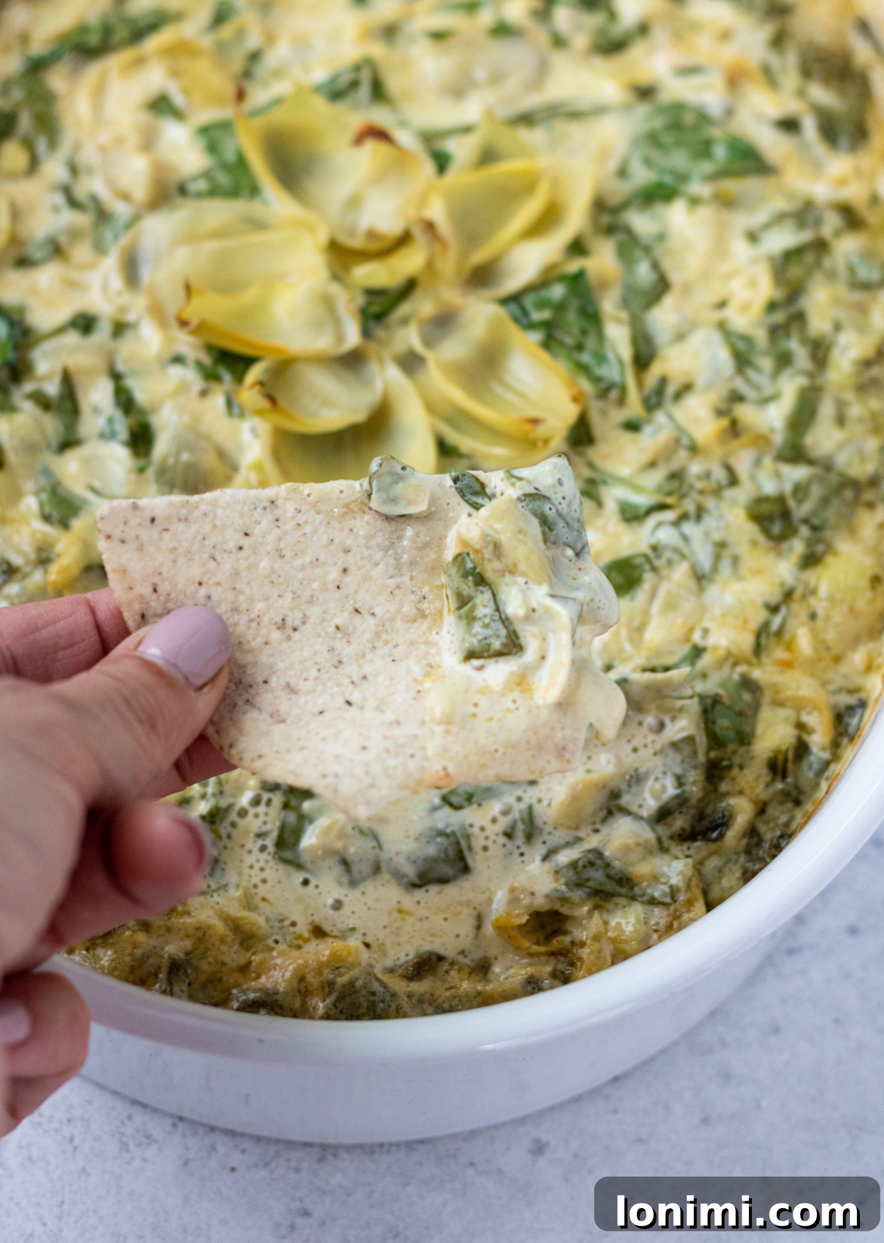 A hand reaching for a scoop of creamy Dairy-Free Spinach Artichoke Dip with a tortilla chip from a casserole dish.