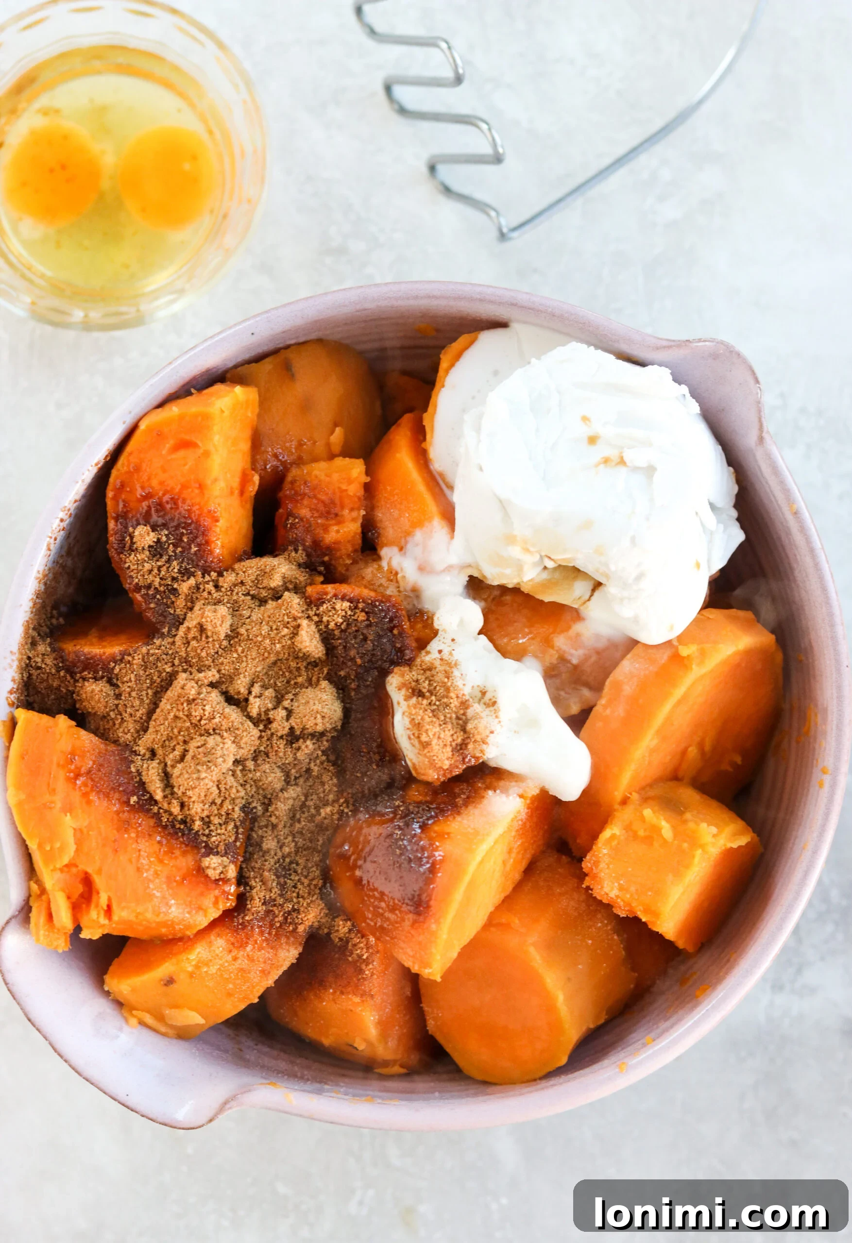 A close-up shot of all ingredients for sweet potato casserole combined in a bowl, with a potato masher and a raw egg visible nearby.