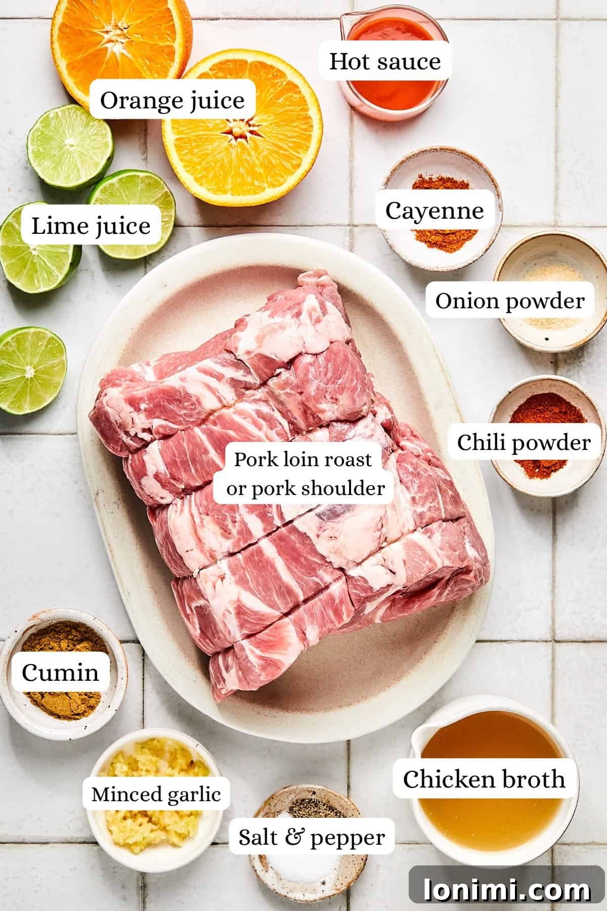 Overhead shot of fresh ingredients for slow cooker pork carnitas including various spices, whole garlic cloves, citrus fruits (lime and orange), a raw pork roast, and a carton of chicken broth.