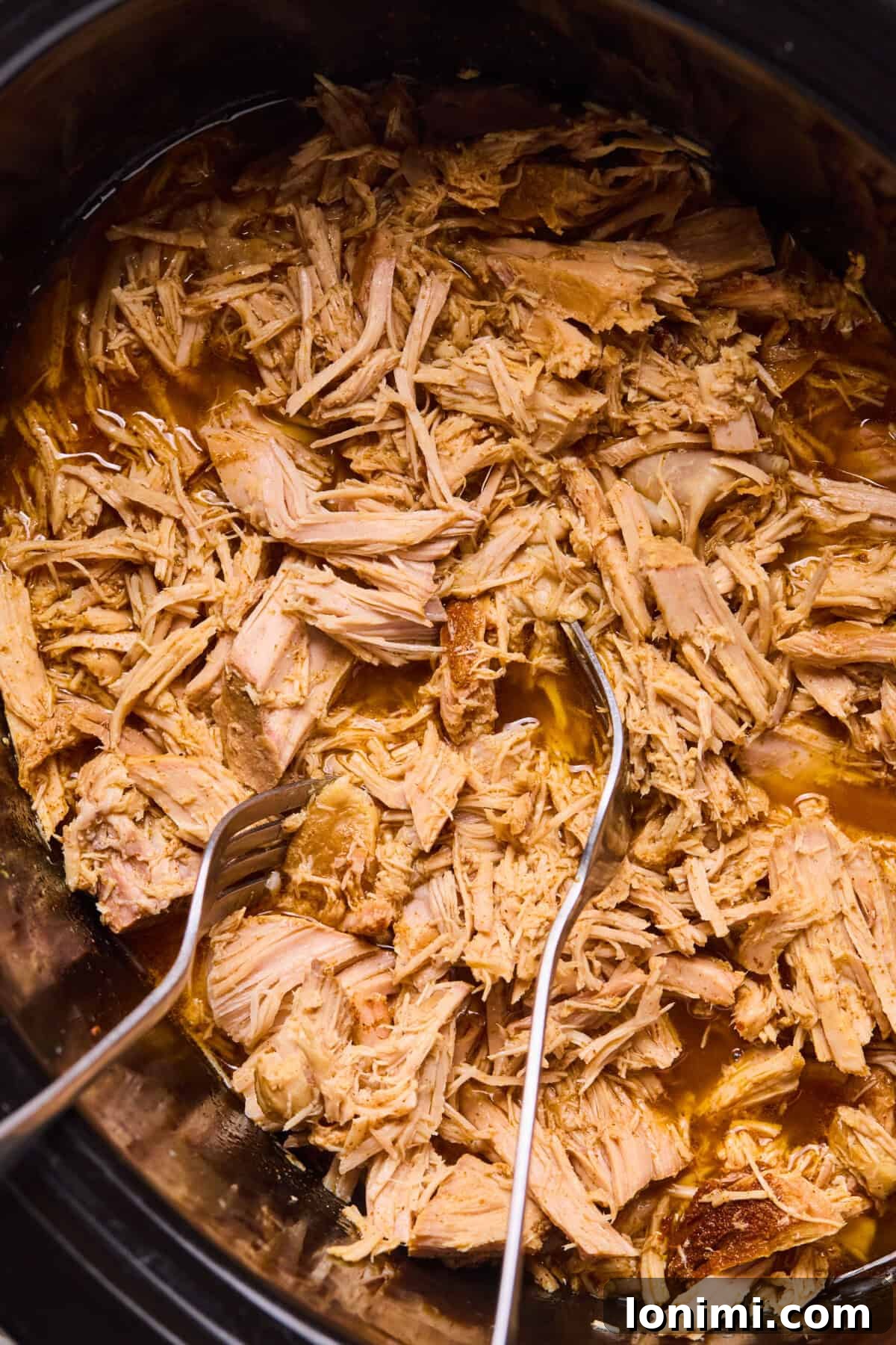 Shredded pork carnitas submerged in their flavorful cooking juices within the slow cooker, absorbing moisture before the final broiling step.