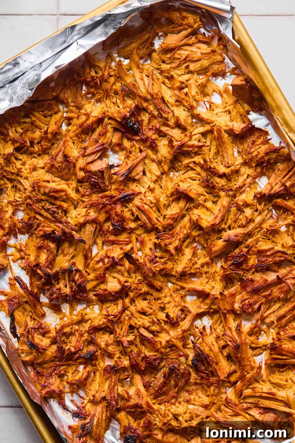 Golden brown and crispy shredded pork carnitas spread out on a foil-lined baking sheet, glistening after being broiled to perfection.