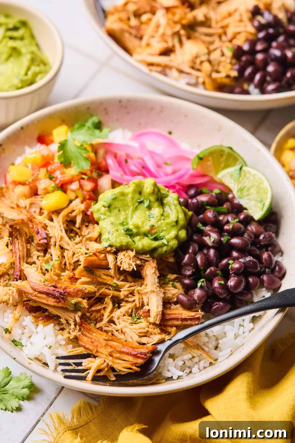 A bowl of slow cooker carnitas surrounded by fresh toppings including creamy guacamole, savory black beans, fluffy rice, tangy pickled onions, and sweet mango salsa, all ready to be assembled into a delicious meal.
