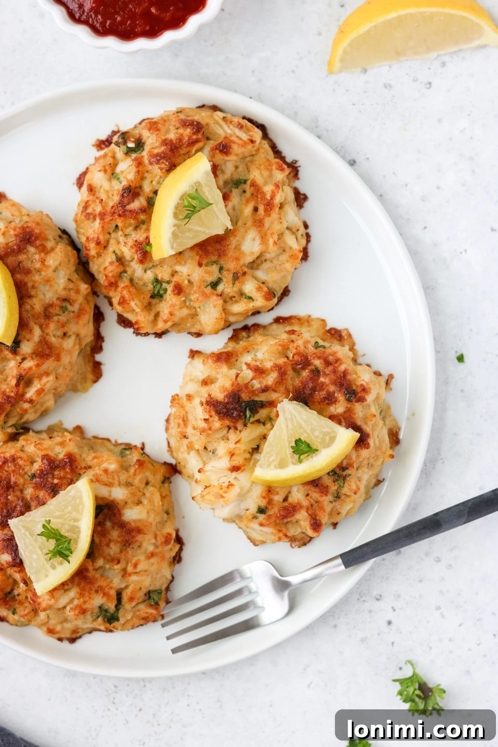Ultimate Gluten Free Maryland Crab Cakes with Delicious Sauce Recipes 3 Four delicious keto crab cakes served on a white plate with a silver fork, garnished with lemon.