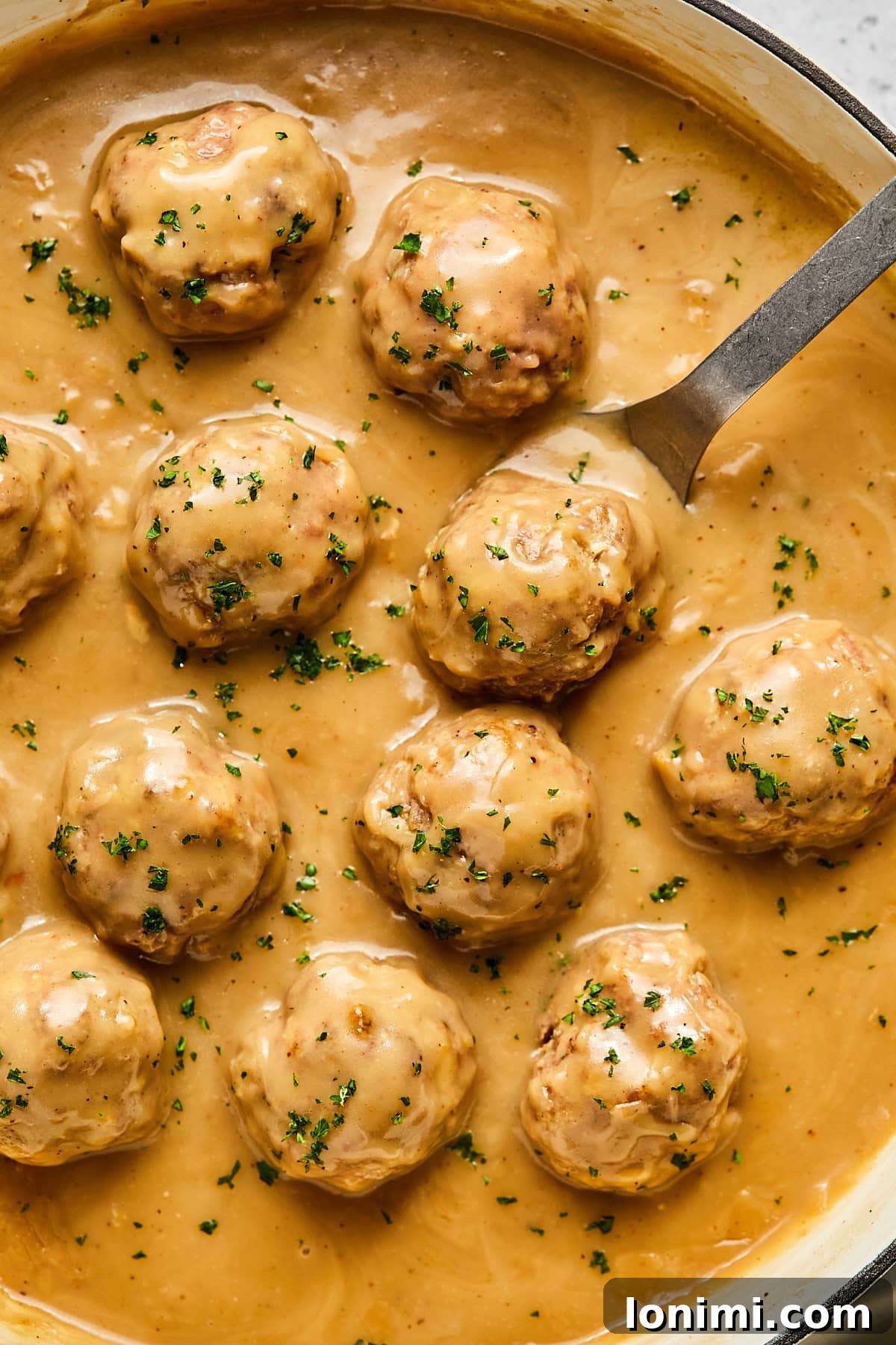 Swedish meatballs generously coated in creamy gravy, served in a pan with a serving spoon, garnished with parsley.