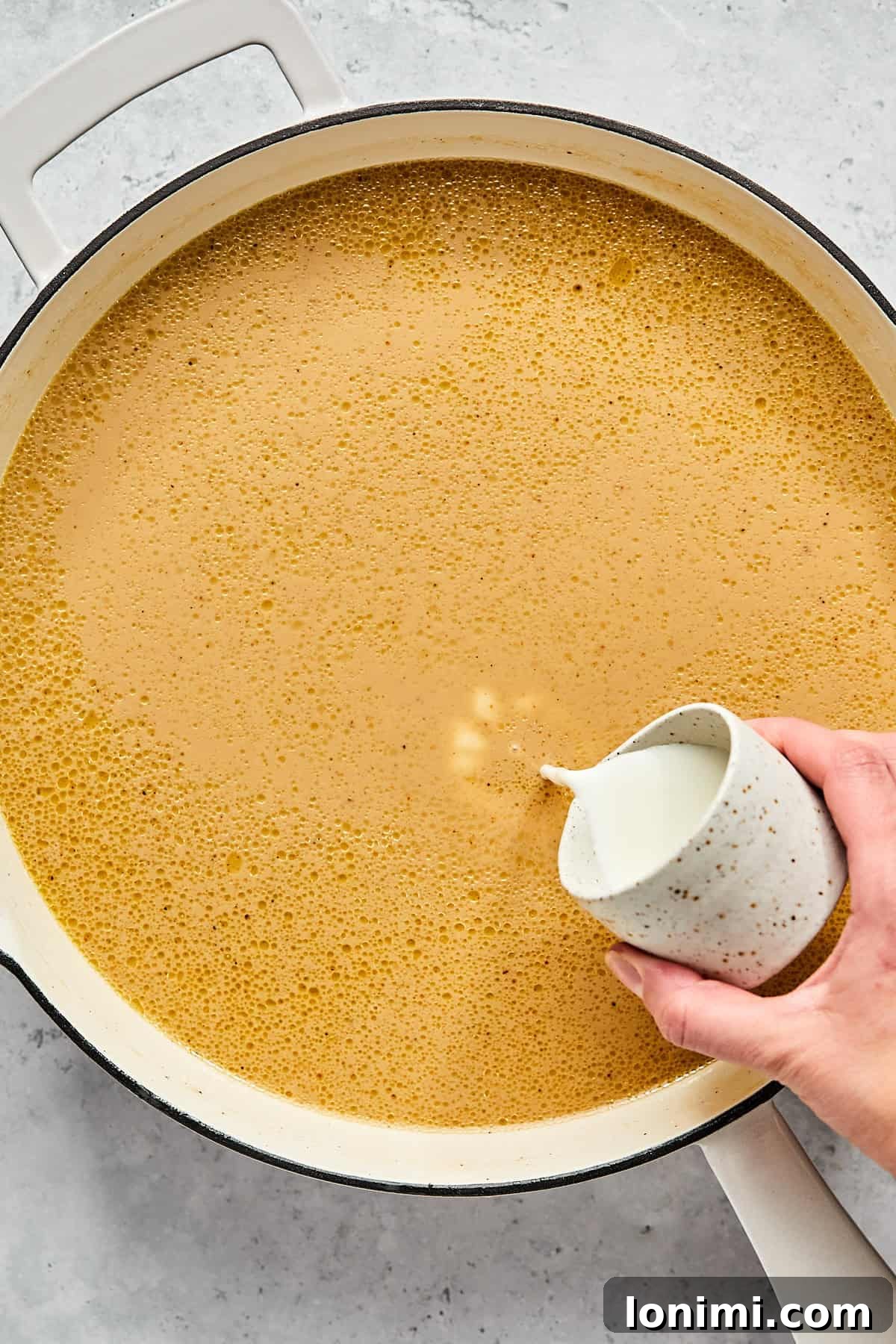 Arrowroot powder slurry being poured into the simmering gravy in a pan, demonstrating the thickening process.