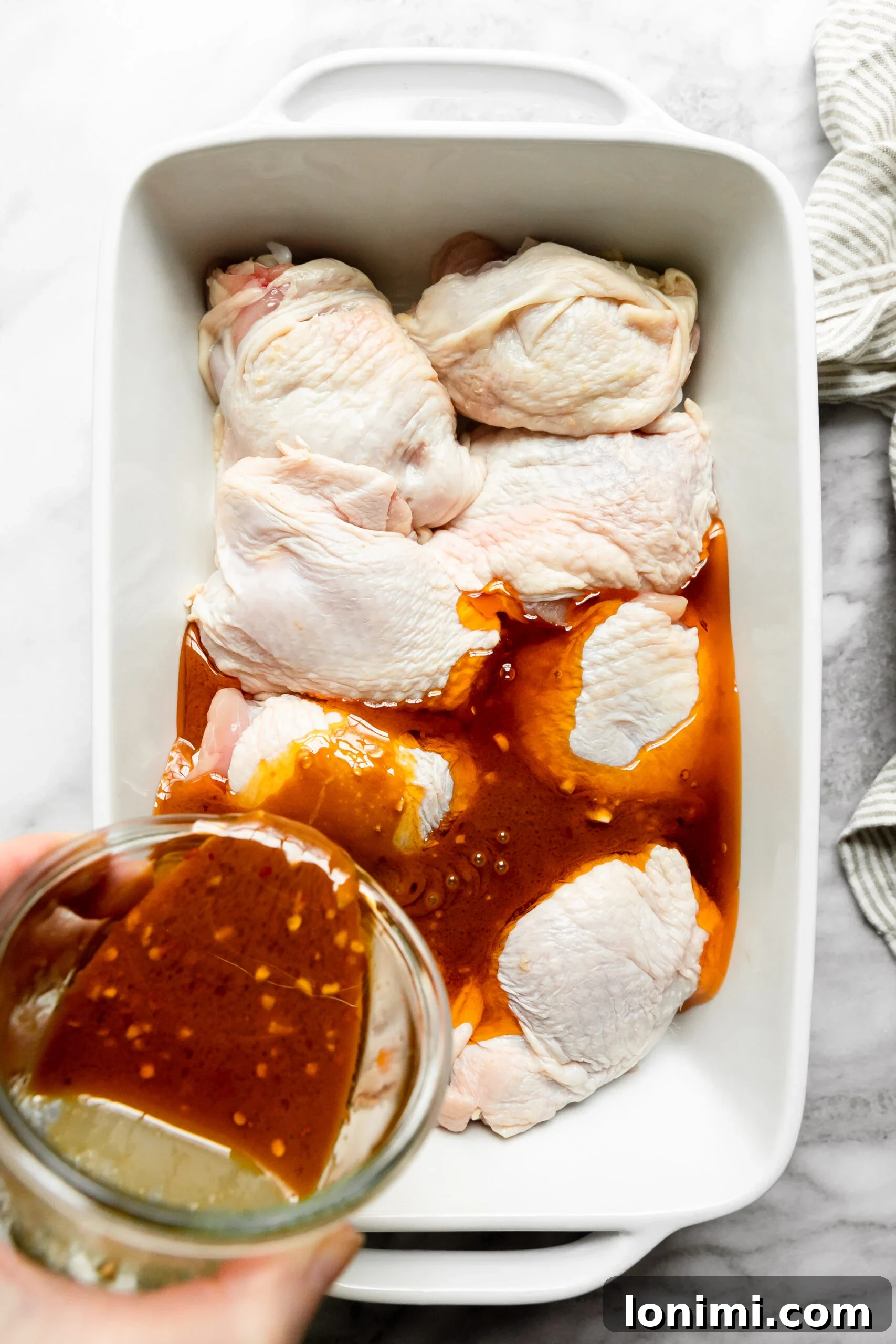Keto Chicken Thighs Perfectly Marinated for Health and Flavor 5 a white casserole dish lined with raw chicken thighs with a glass bowl of marinade pouring the sauce over the top of the chicken