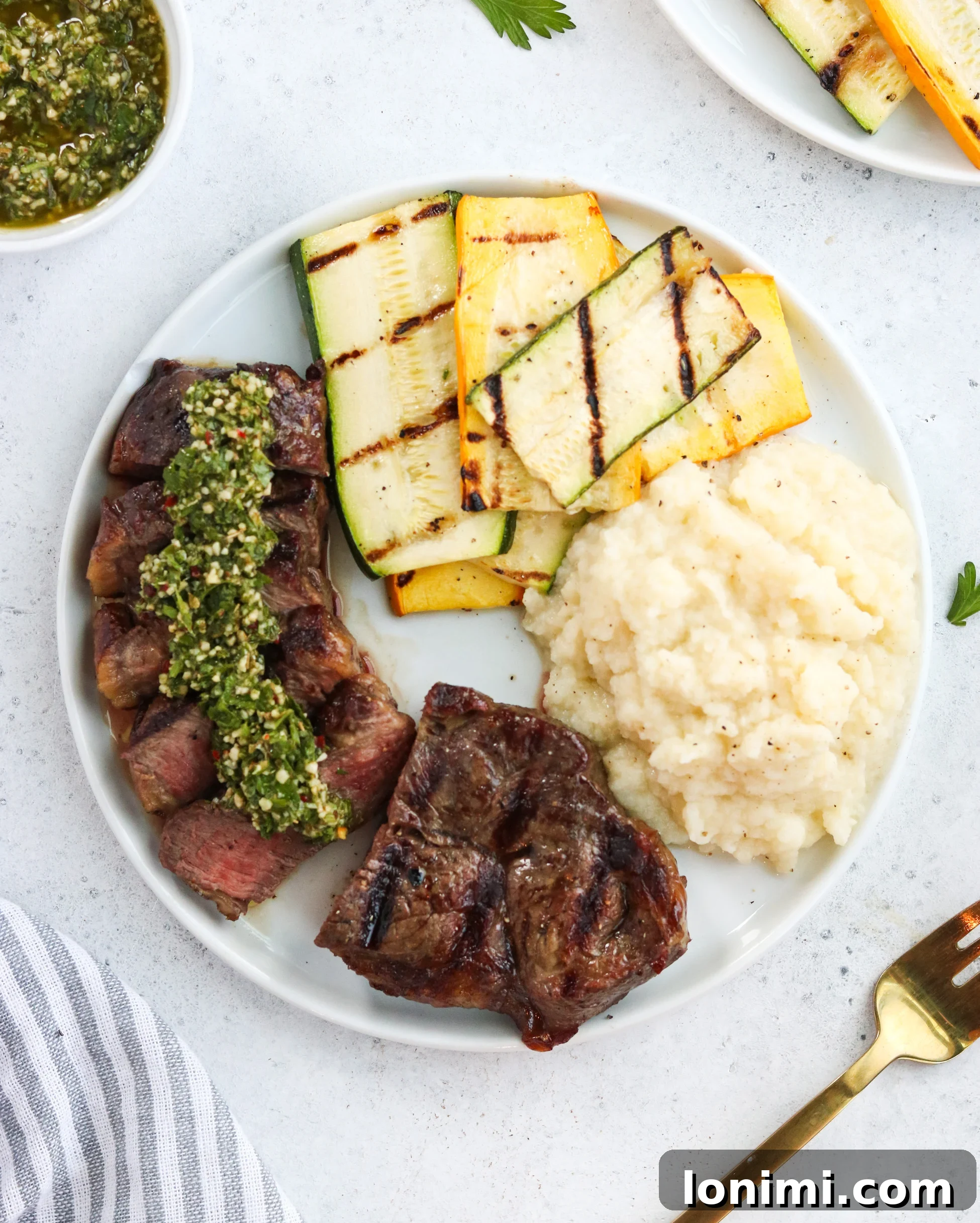 An overhead shot of the complete meal: grilled New York Strip steak topped with chimichurri, surrounded by grilled vegetables and a side dish.