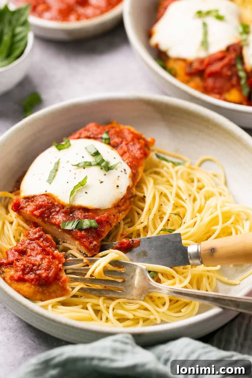 a bowl of spaghetti topped with chicken parmesan being cut with a fork and knife