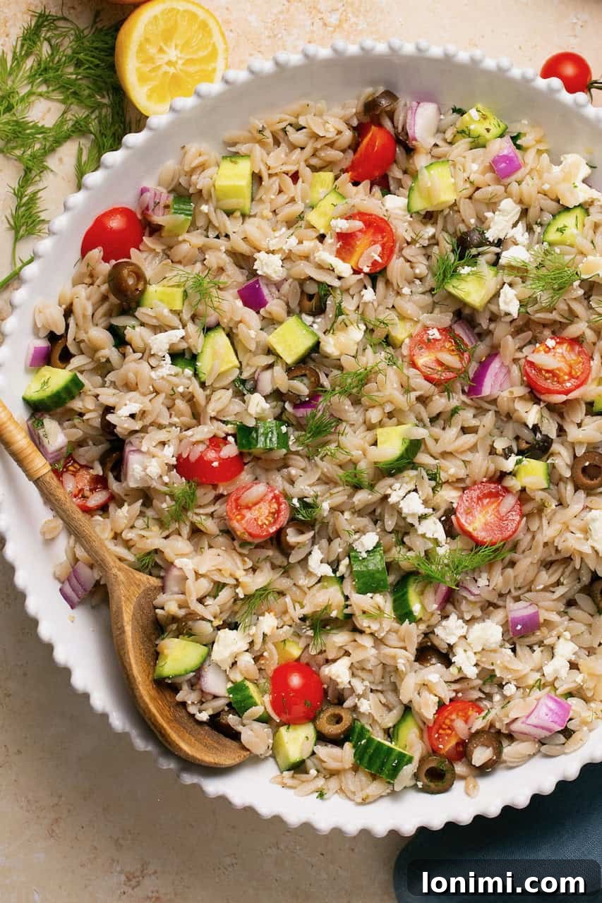 A vibrant gluten-free Mediterranean orzo salad served in a white bowl with a wooden spoon, showcasing fresh vegetables and feta cheese.