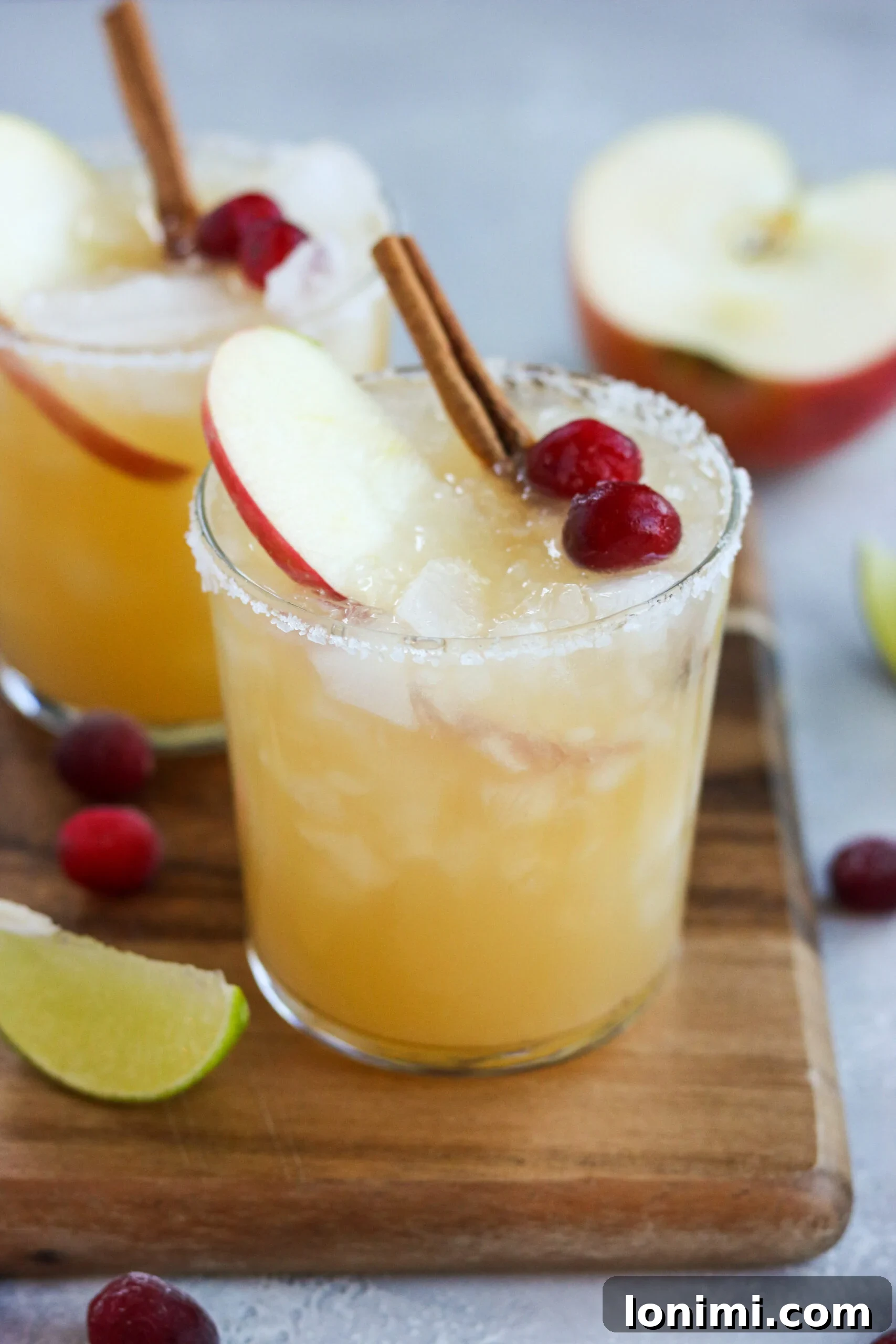 a highball glass filled with ice and an apple cider margarita mixture. Topped with an apple slice, two cranberries and a cinnamon stick with a sea-salt rim