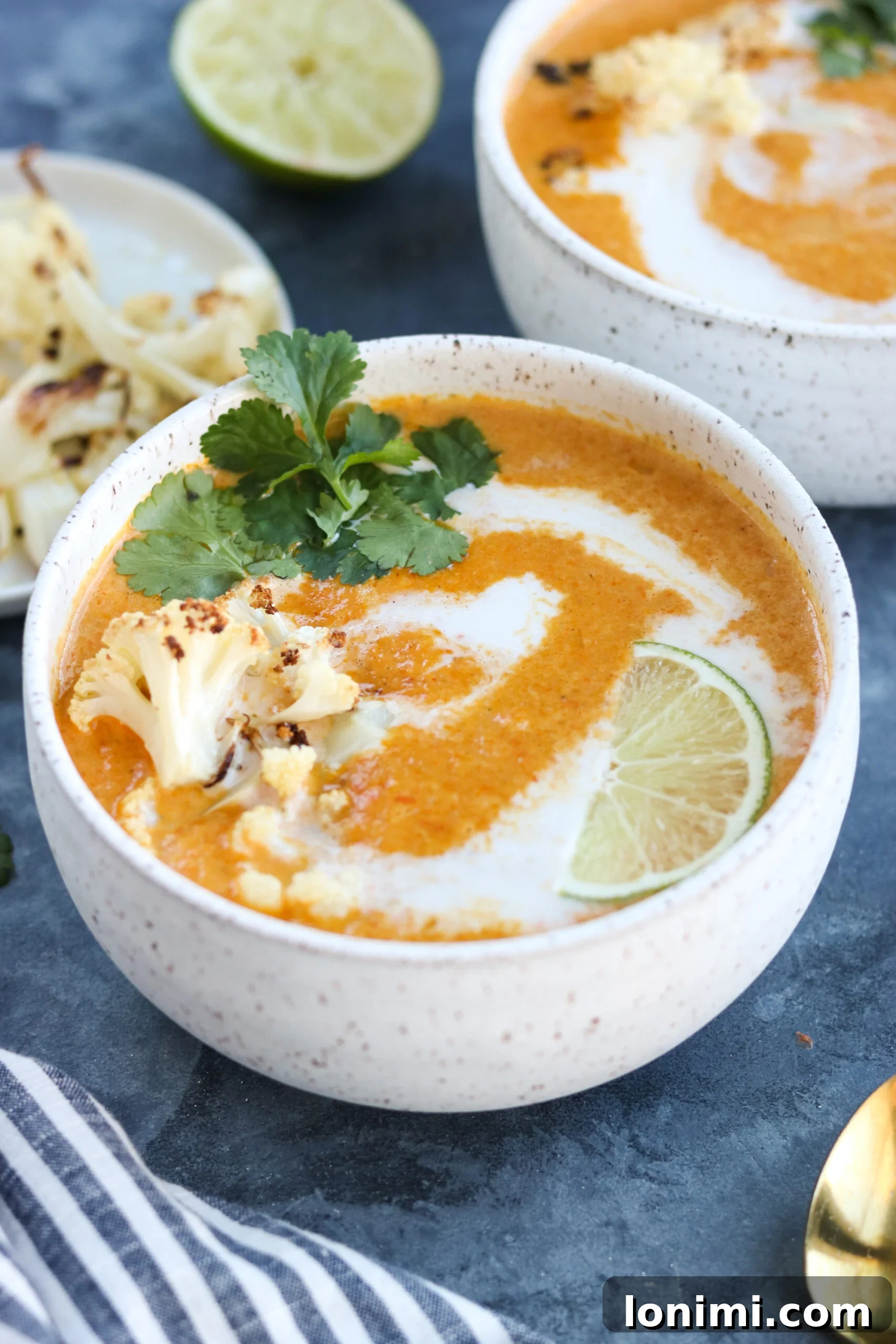 Close-up of Curried Cauliflower Carrot Soup in a white bowl, beautifully garnished with fresh herbs and a lime wedge.