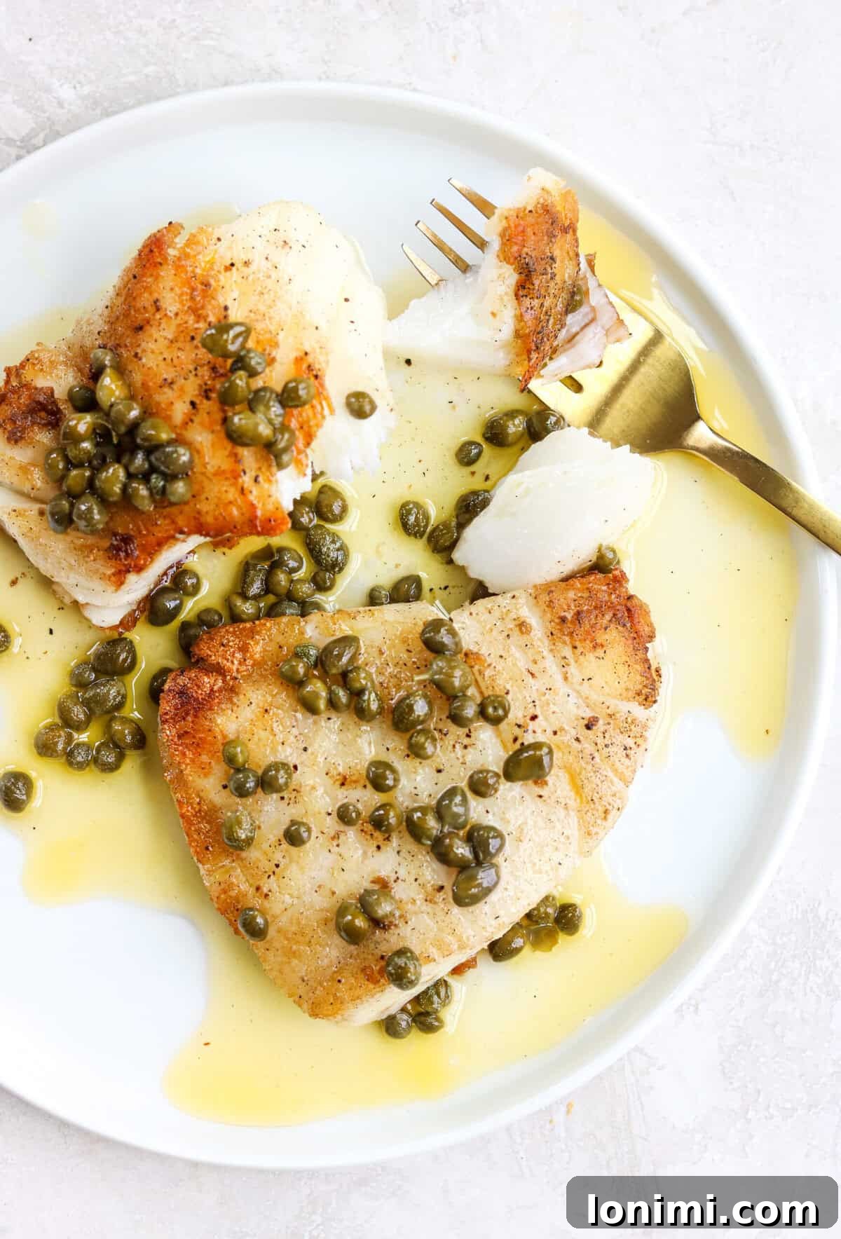 Crispy pan-seared white fish on a white plate with lemon caper sauce and a gold fork with a bit of fish on it, showcasing the exquisite texture and vibrant sauce.