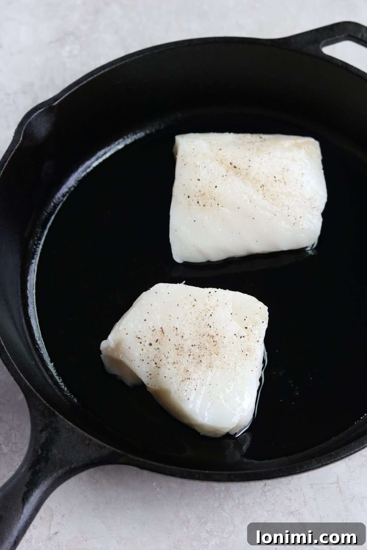 Two sea bass fillets sizzling in a hot cast iron pan, skin-side down, developing a golden crust.