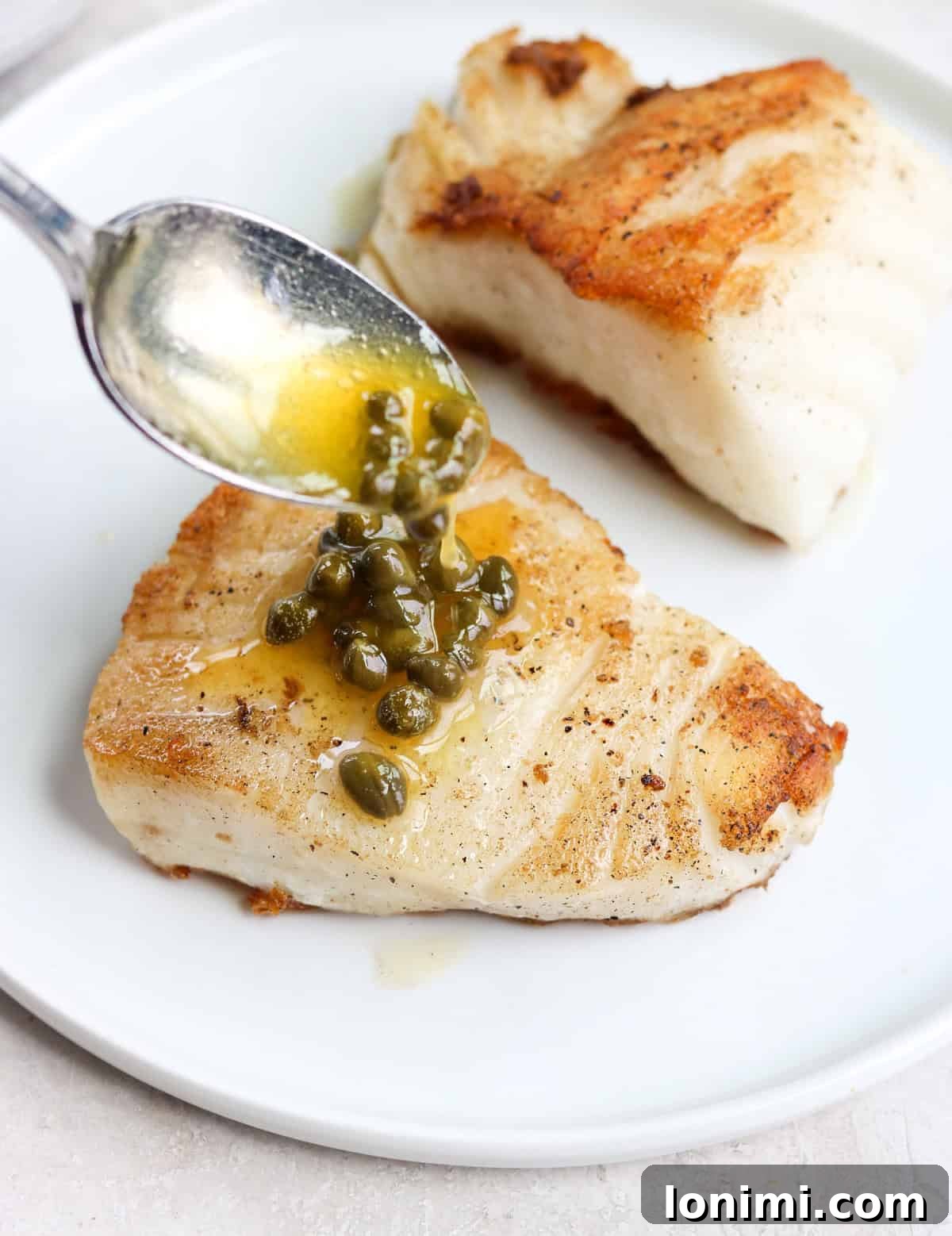 A silver spoon drizzling vibrant lemon caper butter over a perfectly cooked piece of Chilean sea bass on a white plate, garnished with fresh parsley.