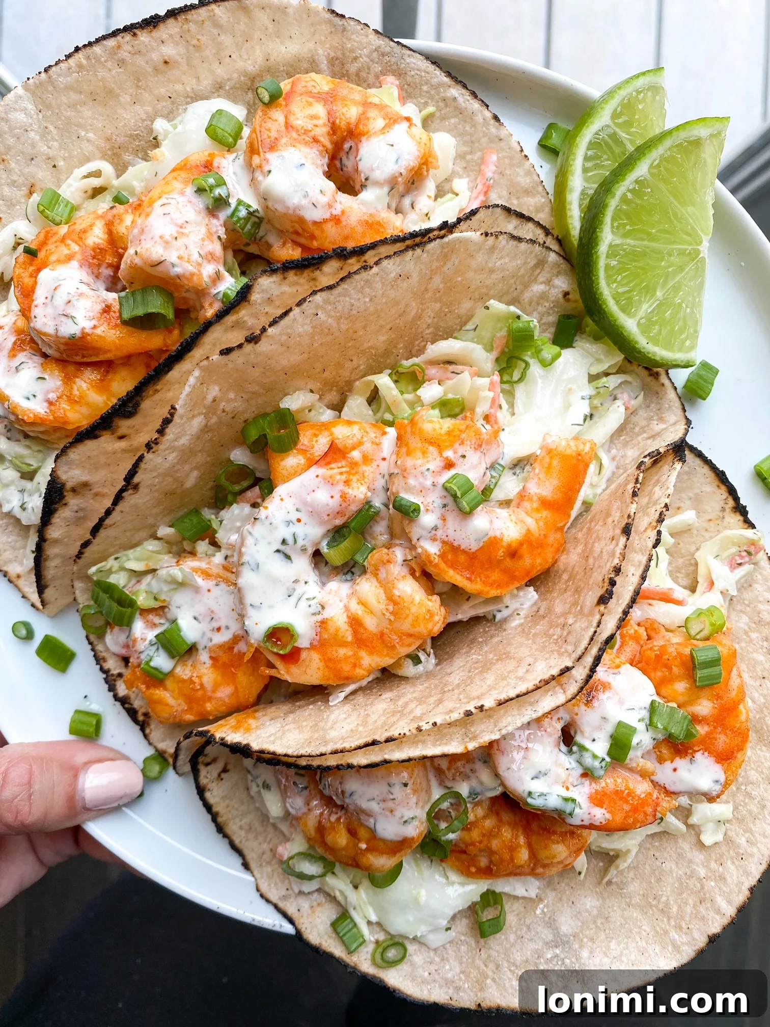 Tacos on a plate ready to eat A hand holding a white plate showcasing a trio of exquisitely prepared Paleo Buffalo Shrimp Tacos, garnished with fresh cilantro, ready to be enjoyed.