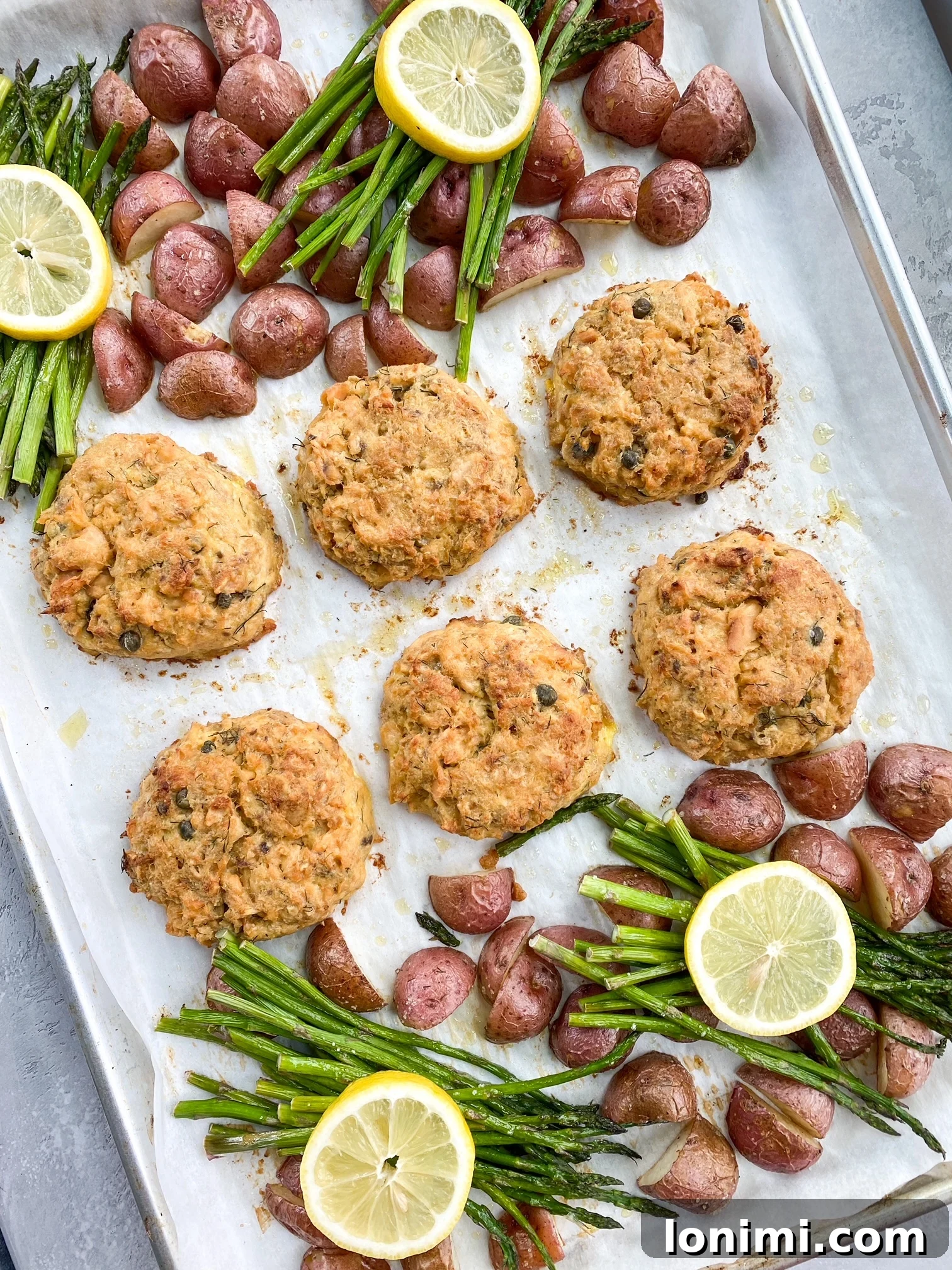 Lemon Caper Salmon Cakes in a Sheet Pan Freshly prepared Lemon Caper Salmon Cakes baking on a sheet pan alongside golden baby potatoes and green asparagus tips.