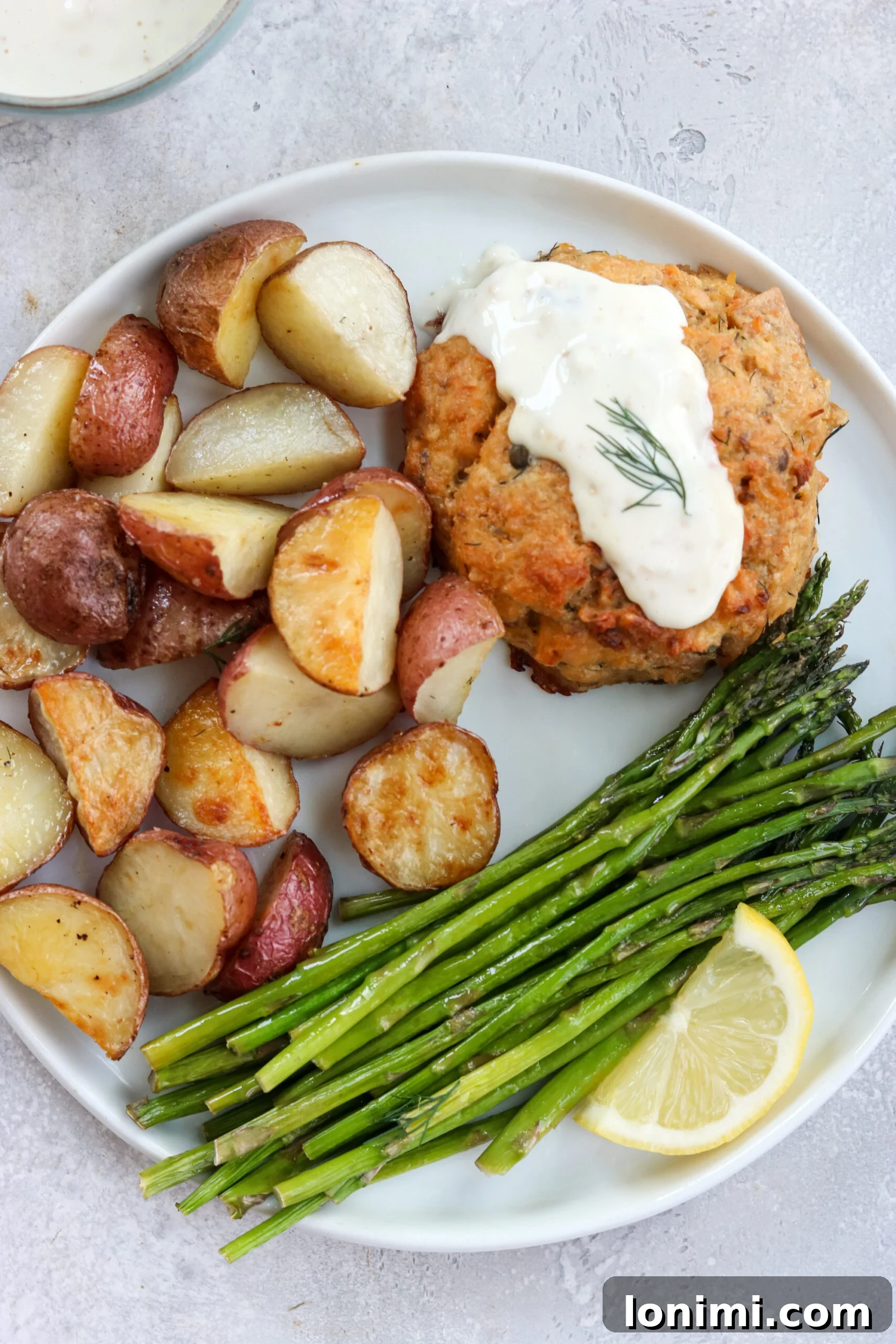 cooked salmon cake and vegetables A close-up of a perfectly cooked Lemon Caper Salmon Cake on a white plate, served with roasted potatoes and asparagus, and a dollop of lemon-garlic aioli.