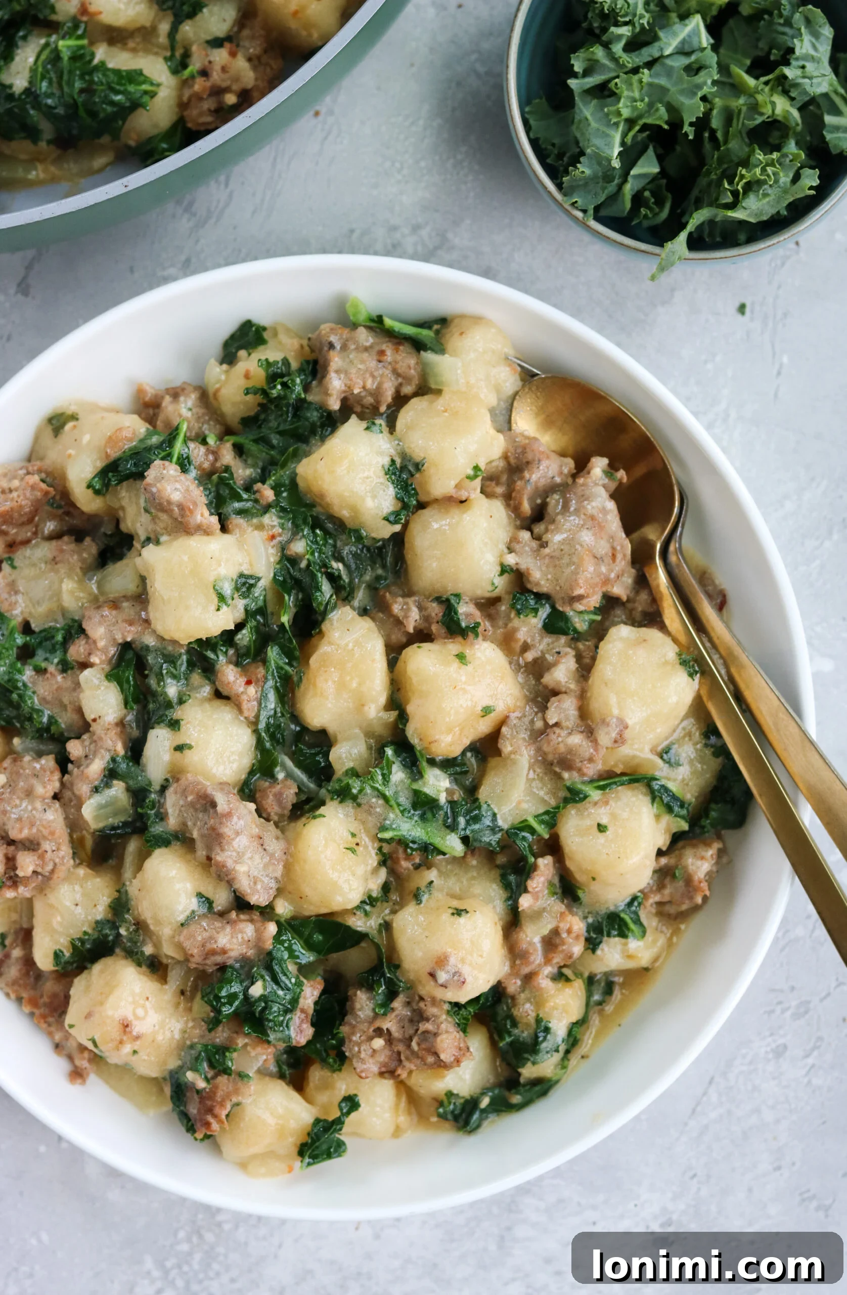 A close-up shot of Paleo Sausage Kale Gnocchi in a white bowl, with a large skillet of the dish in the background, highlighting the fresh ingredients and inviting texture.
