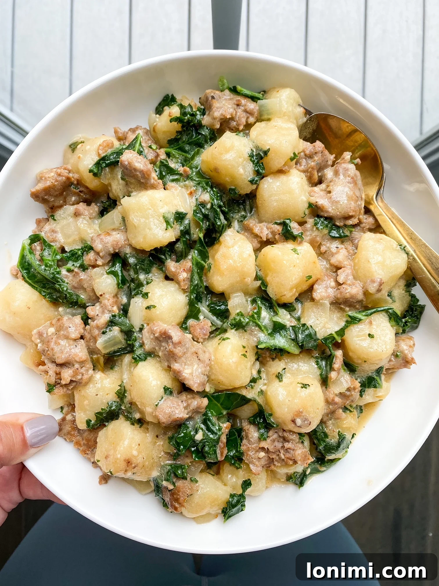 A hand holding a white bowl filled with a generous serving of Paleo Sausage Kale Gnocchi, showcasing the rich, creamy texture and vibrant ingredients.