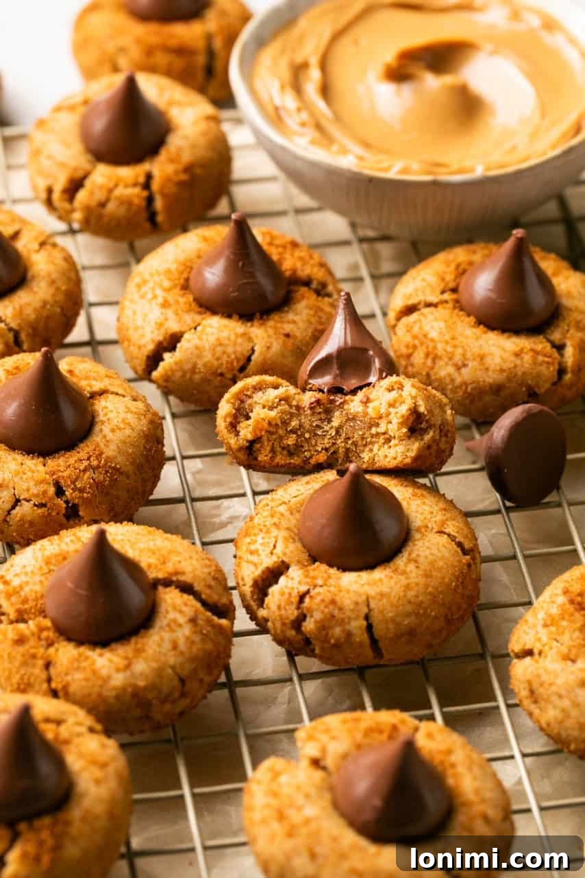 Foolproof Gluten Free Peanut Butter Blossoms 2 gluten free peanut butter blossoms on a cooling rack with a bowl of peanut butter in the background