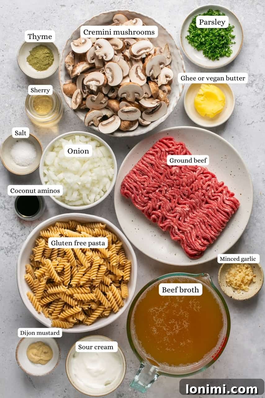 A collection of fresh ingredients laid out, including cremini mushrooms, ground beef, onions, garlic, pasta, and herbs, for making gluten-free beef stroganoff.
