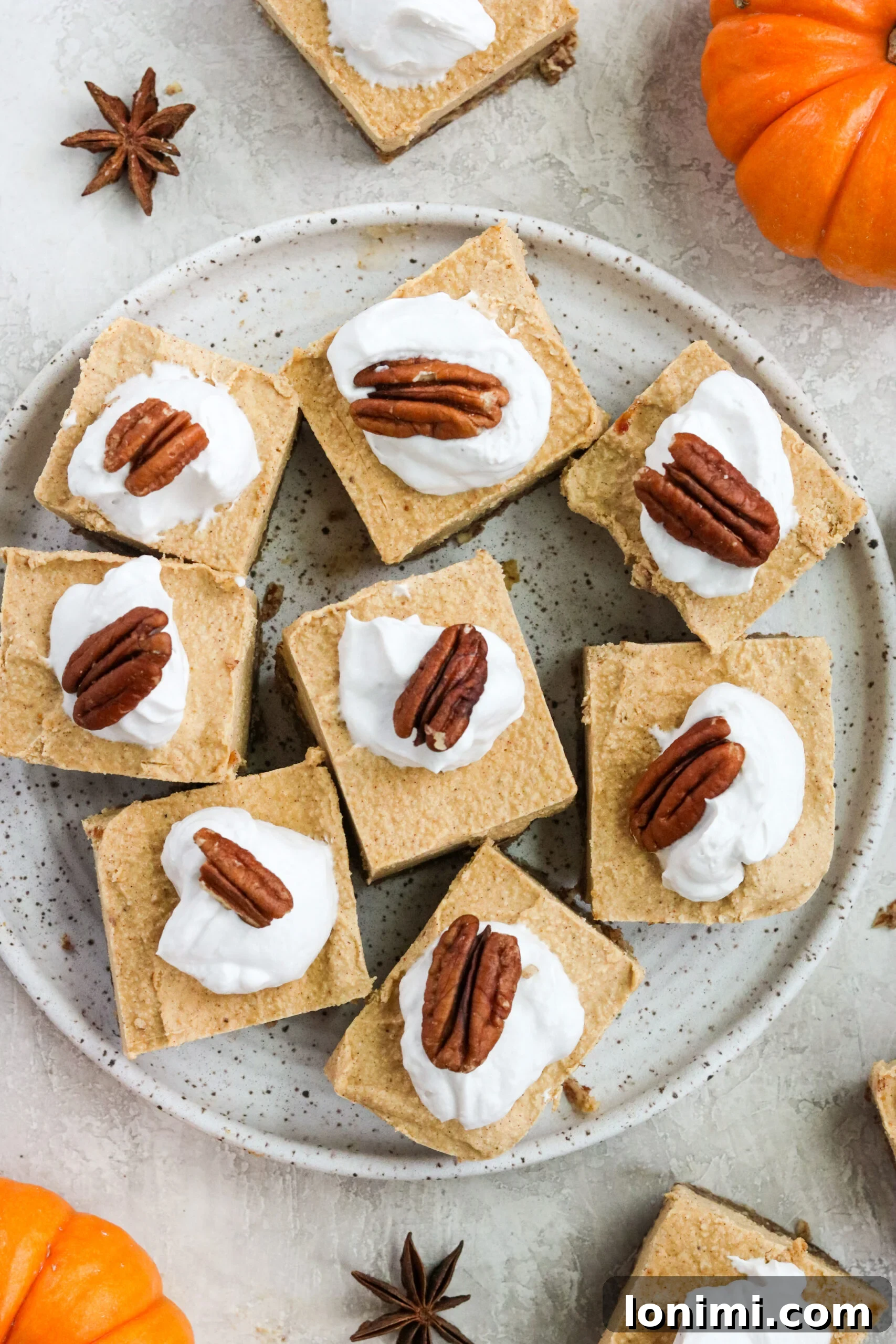 seven pumpkin cheesecake bars topped with dairy free whipped cream and pecans served on a speckled white plate