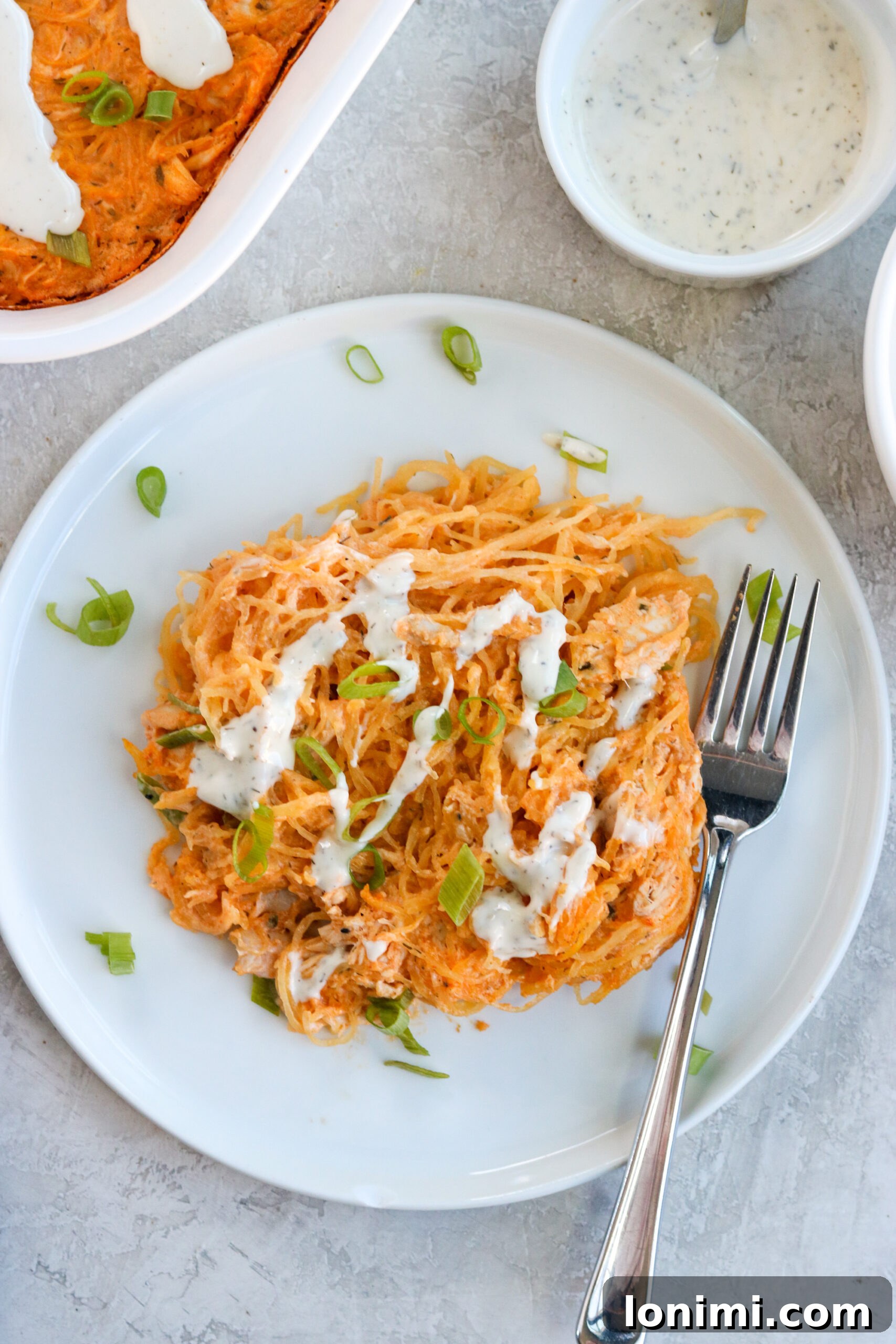 Guilt-Free Buffalo Chicken Spaghetti Squash Bake 5 A generous serving of healthy Buffalo Chicken Spaghetti Squash Casserole on a white plate, elegantly drizzled with dairy-free ranch dressing and garnished with vibrant diced green onions, showcasing its appetizing appeal.