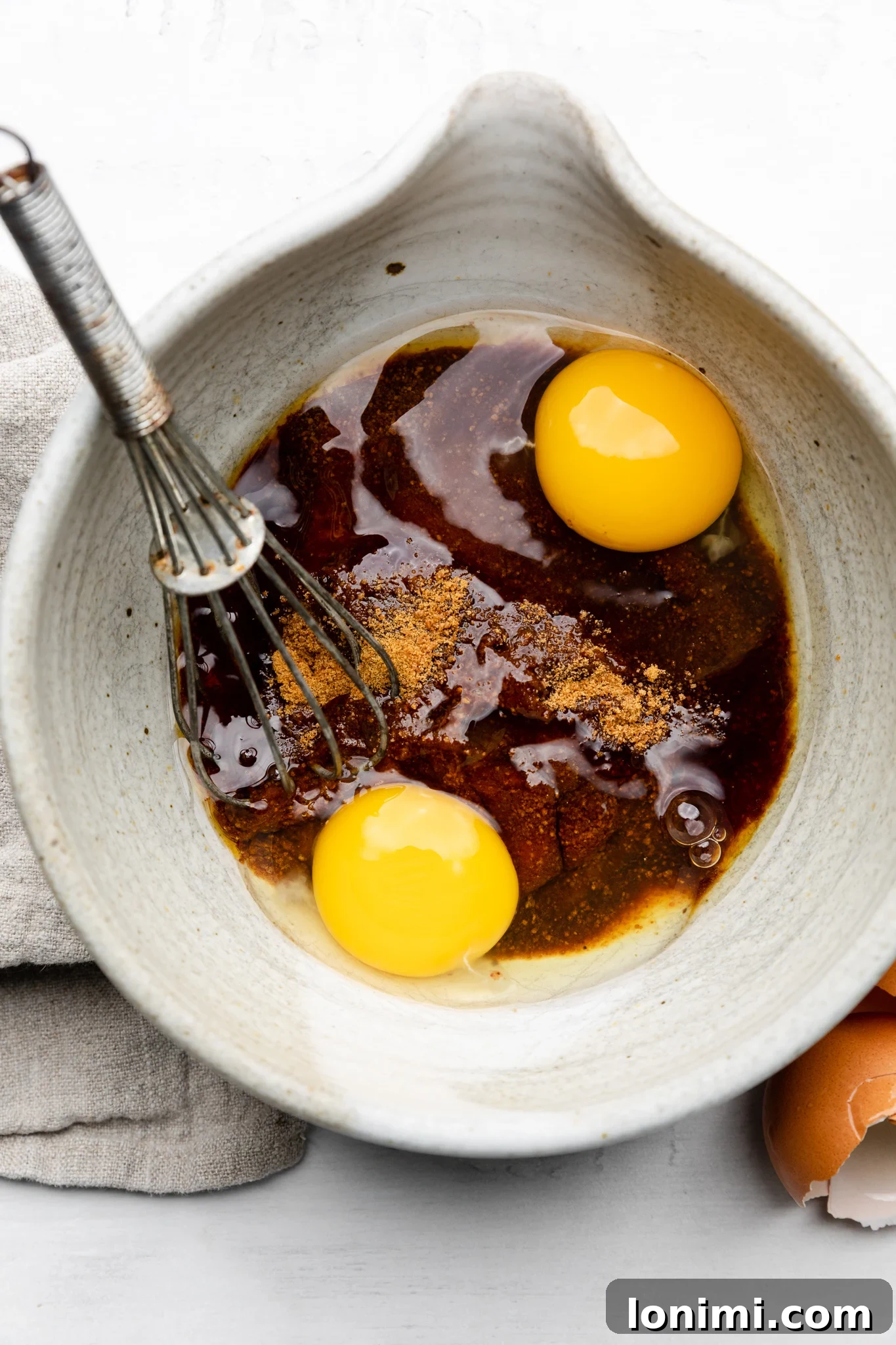 A close-up shot of eggs, maple syrup, coconut sugar, and vanilla extract being whisked together in a white mixing bowl with a metal whisk, illustrating the wet ingredient preparation for the mini chocolate cake recipe.