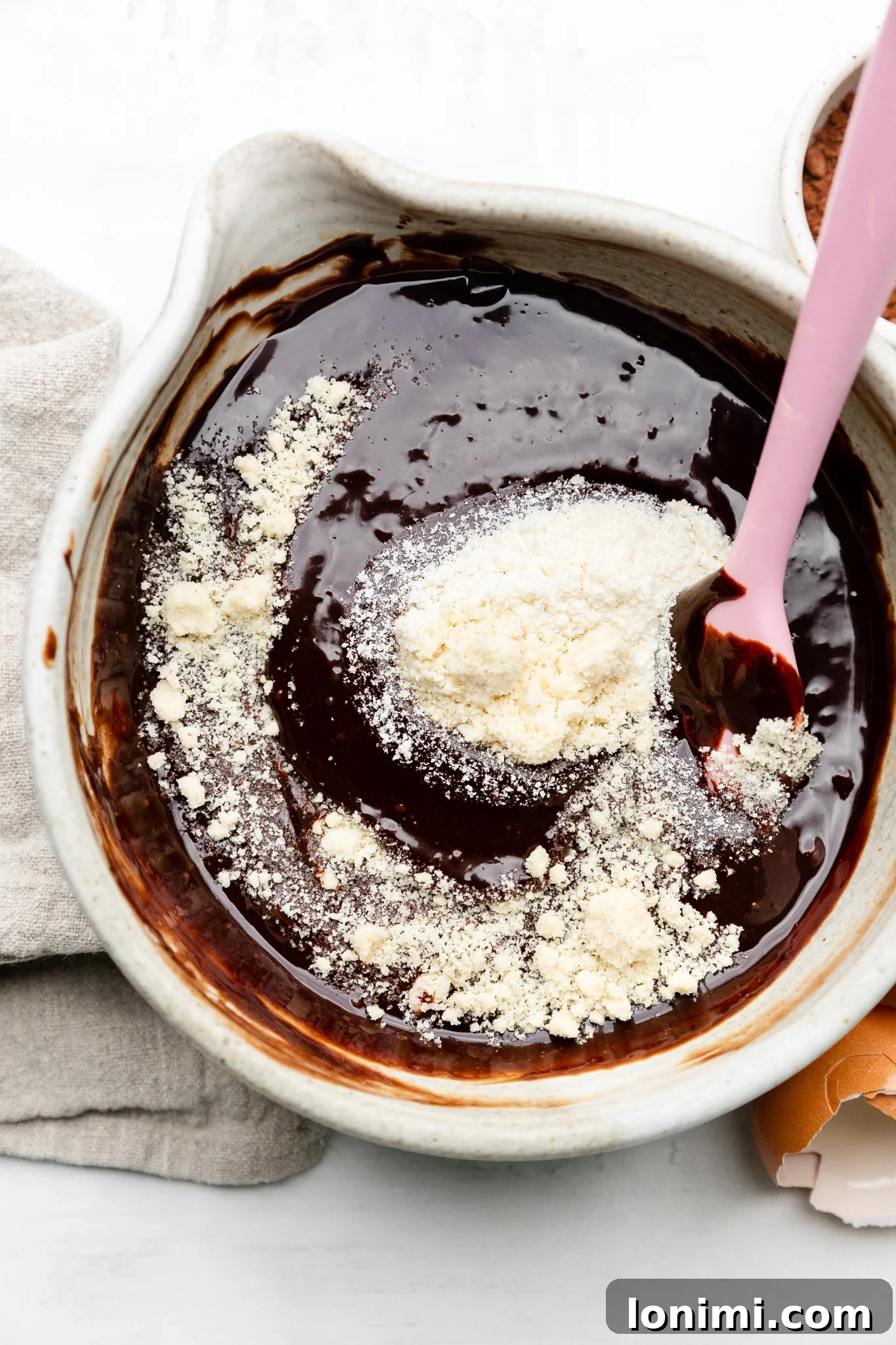 A mixing bowl containing melted dark chocolate, almond flour, and coconut flour, with a pink spatula gently folding the ingredients, demonstrating the final batter preparation for the mini chocolate cake recipe.