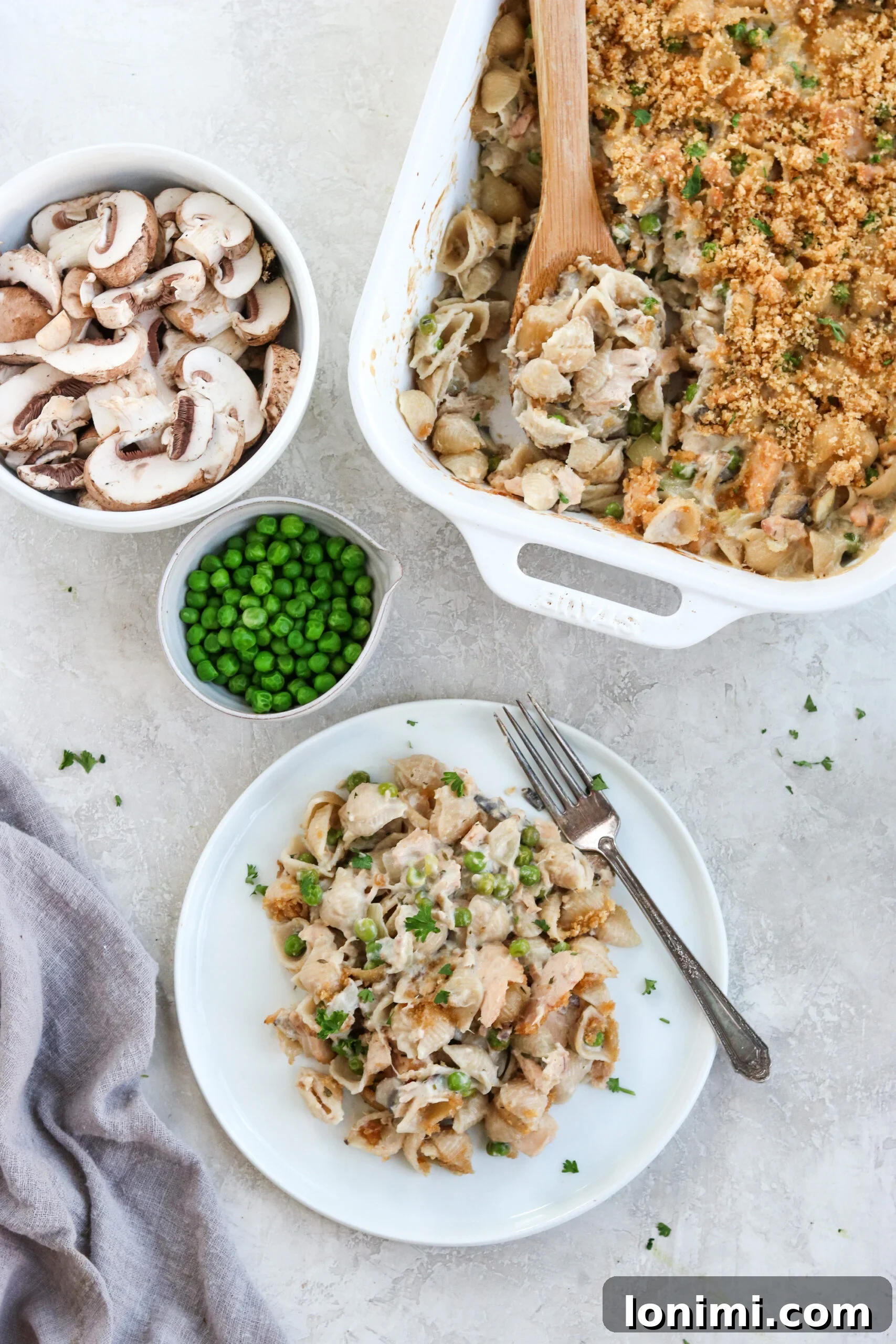 Wholesome Tuna Noodle Bake 5 Tuna noodle casserole served on a white plate with the white casserole dish and a bowl of mushrooms and peas on the side, garnished with fresh herbs.