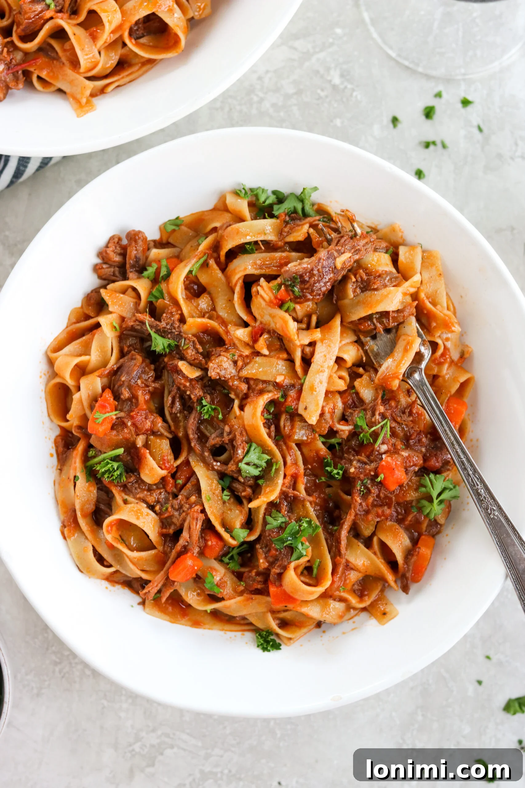 Tender Beef Short Rib Ragu in a white bowl, garnished with fresh parsley, with a fork digging in.