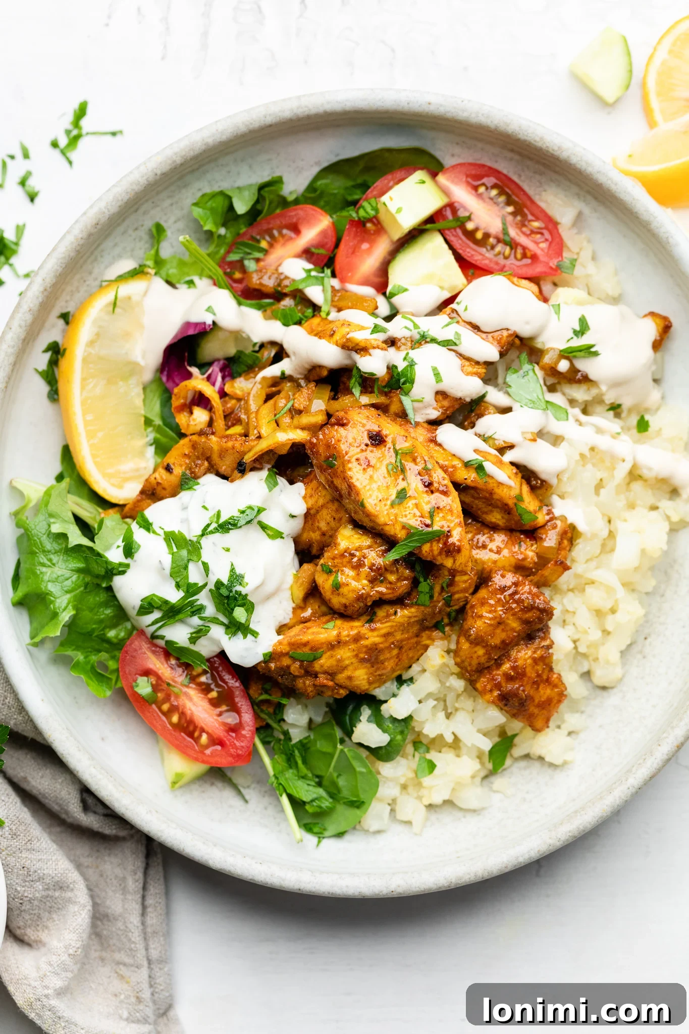 A beautifully arranged Sheet Pan Chicken Shawarma Bowl featuring succulent chicken, fluffy cauliflower rice, vibrant tomatoes, fresh parsley, a wedge of lemon, and generous dollops of tzatziki and lemon tahini dressing.
