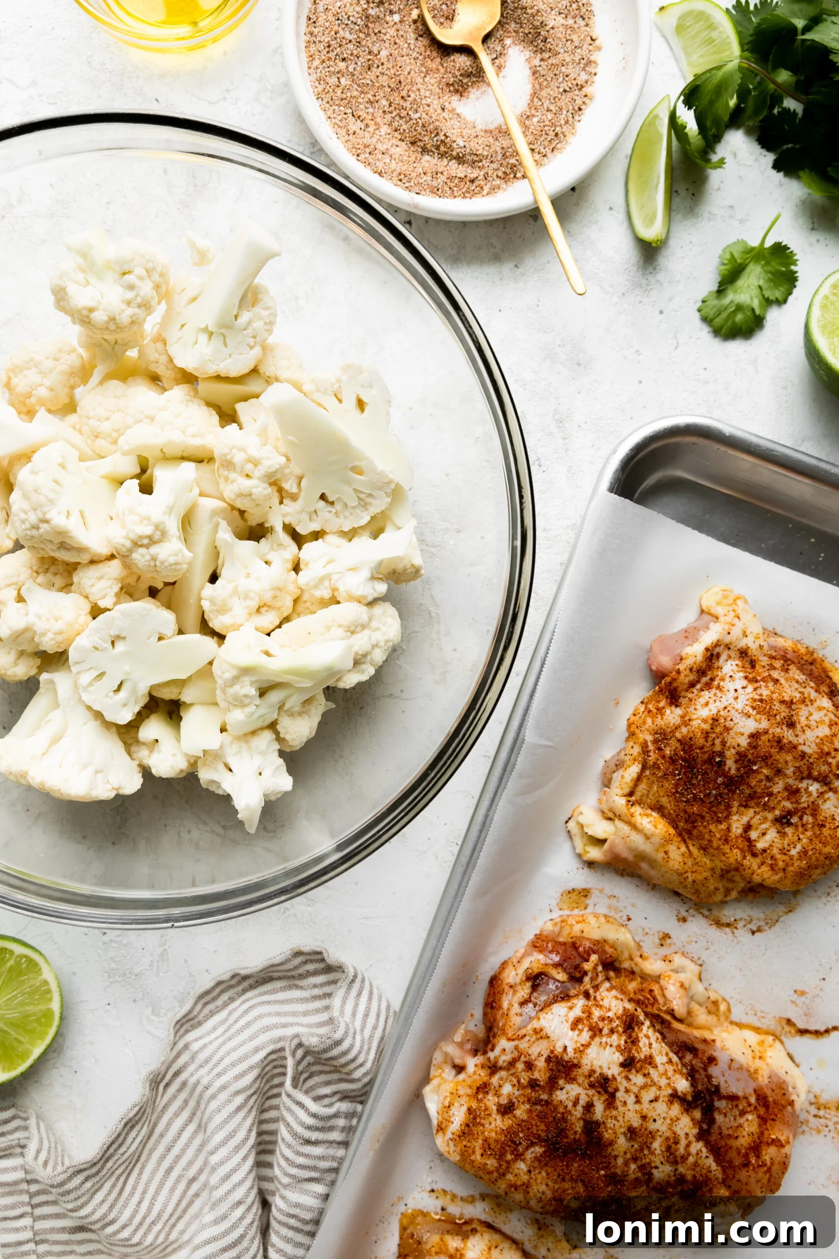Sheet Pan Fiesta Bowls with Chicken and Roasted Cauliflower 4 a close up photo of raw cauliflower in a glass bowl next to chicken thighs on a silver baking pan with a bowl of cilantro lime sauce next to it.