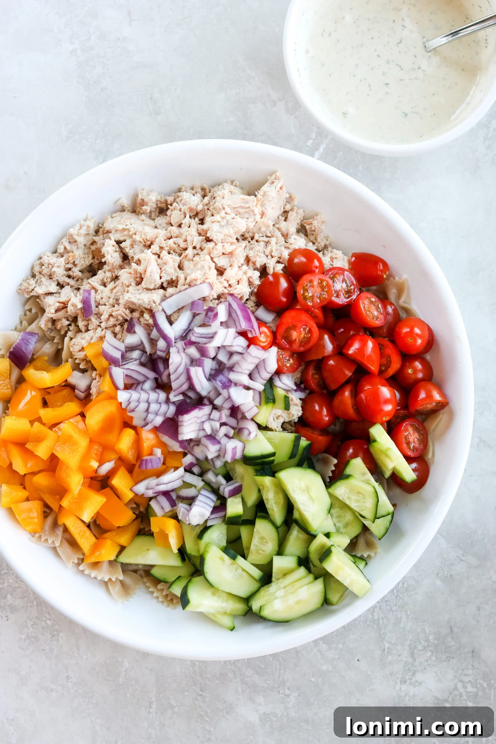 Diced vegetables for pasta salad in a white bowl next to a bowl of creamy mayo sauce.