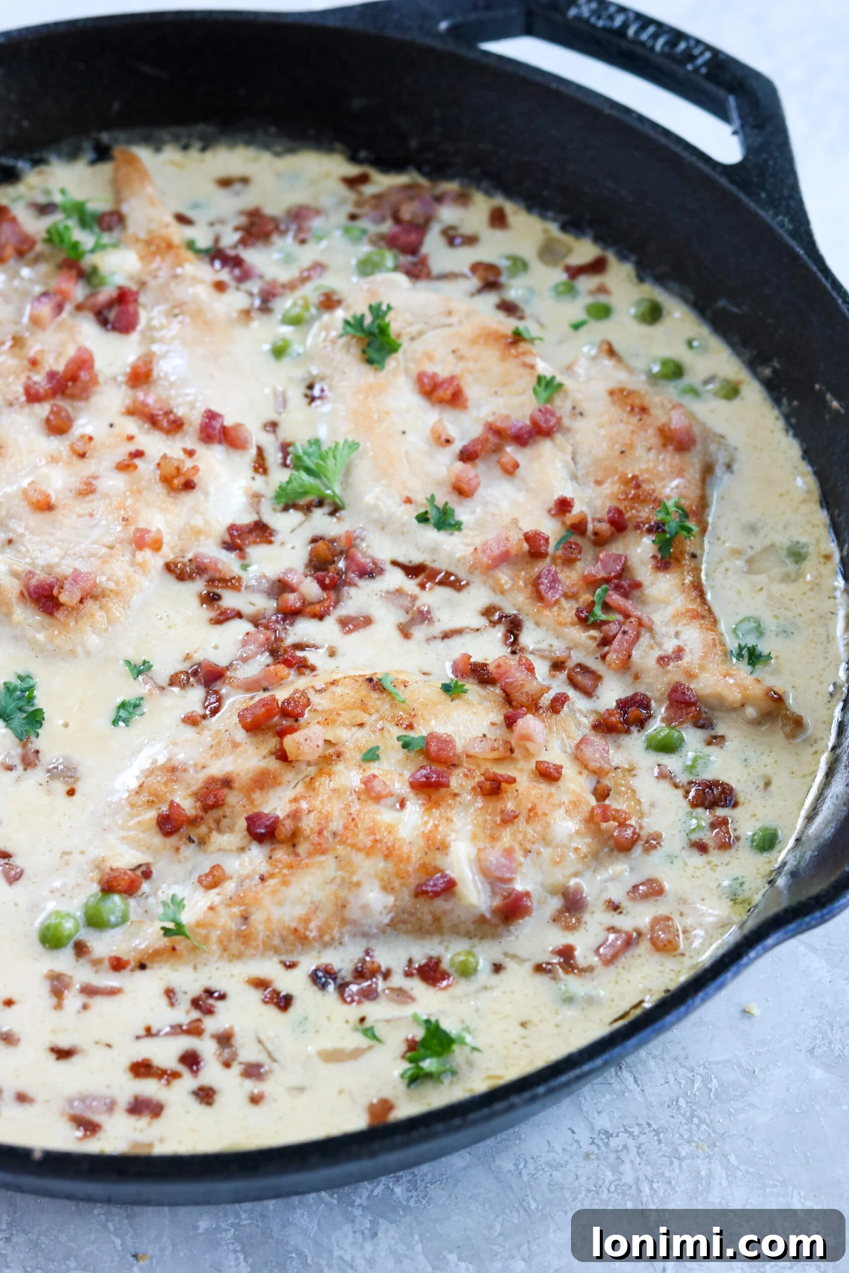 A close-up view of a cast iron pan brimming with perfectly cooked chicken breasts, vibrant green peas, and a luxurious lemon cream sauce, generously topped with crisp pancetta and fresh parsley.