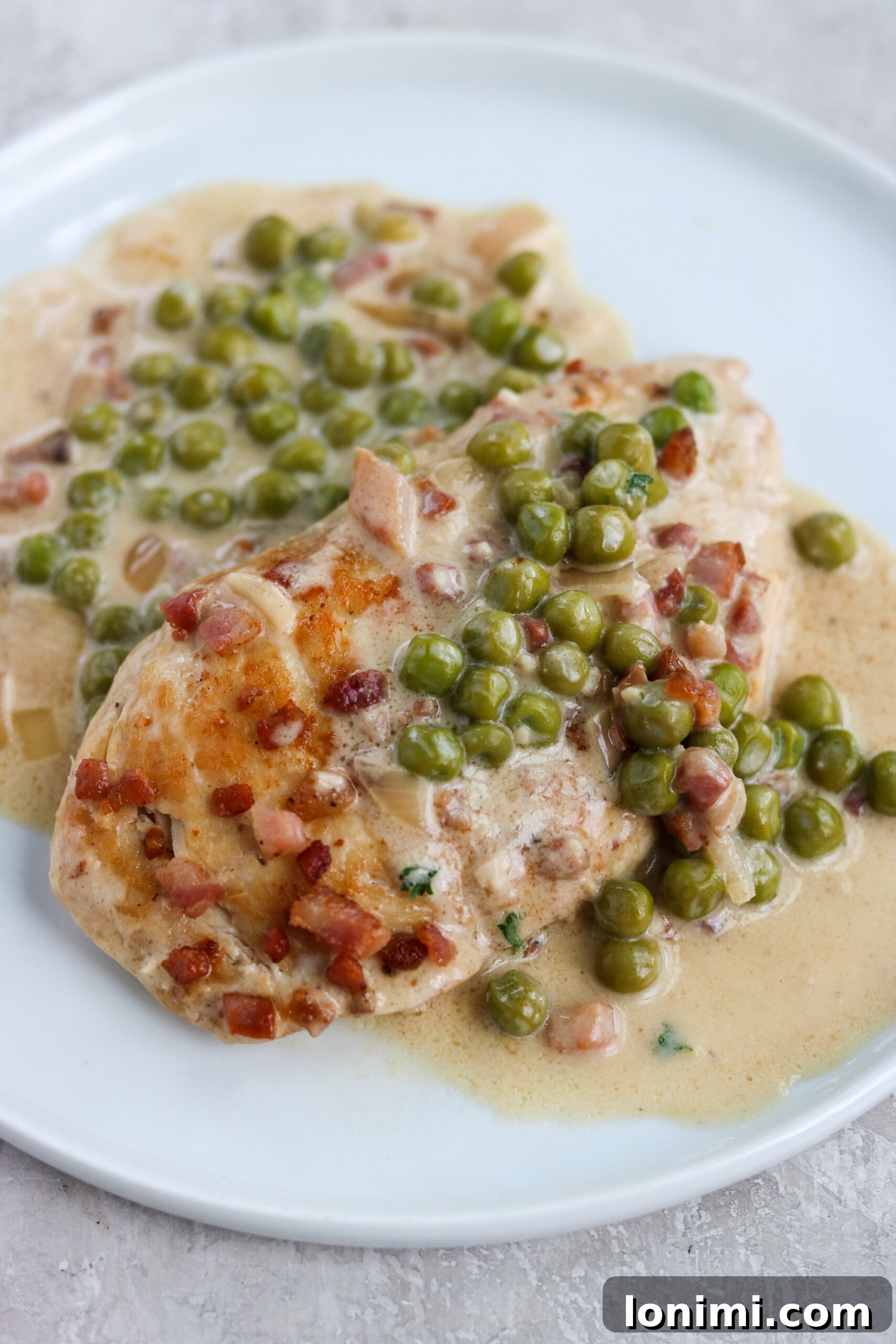 A close-up photo showcasing a perfectly golden-brown cooked chicken breast, generously coated in the vibrant lemon cream sauce with tender green peas and topped with crispy pancetta.