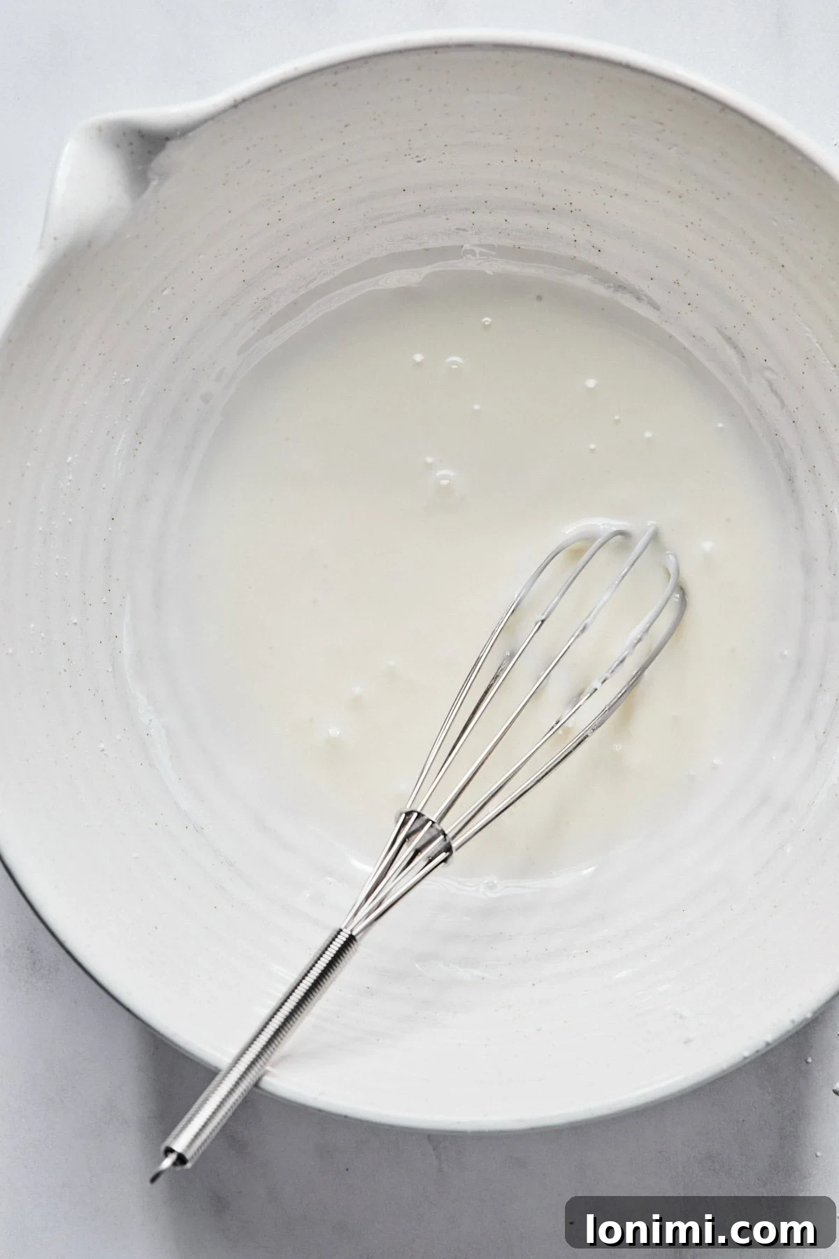 Smooth almond glaze in a medium white bowl with a whisk, ready to be applied to the cooled cookies.