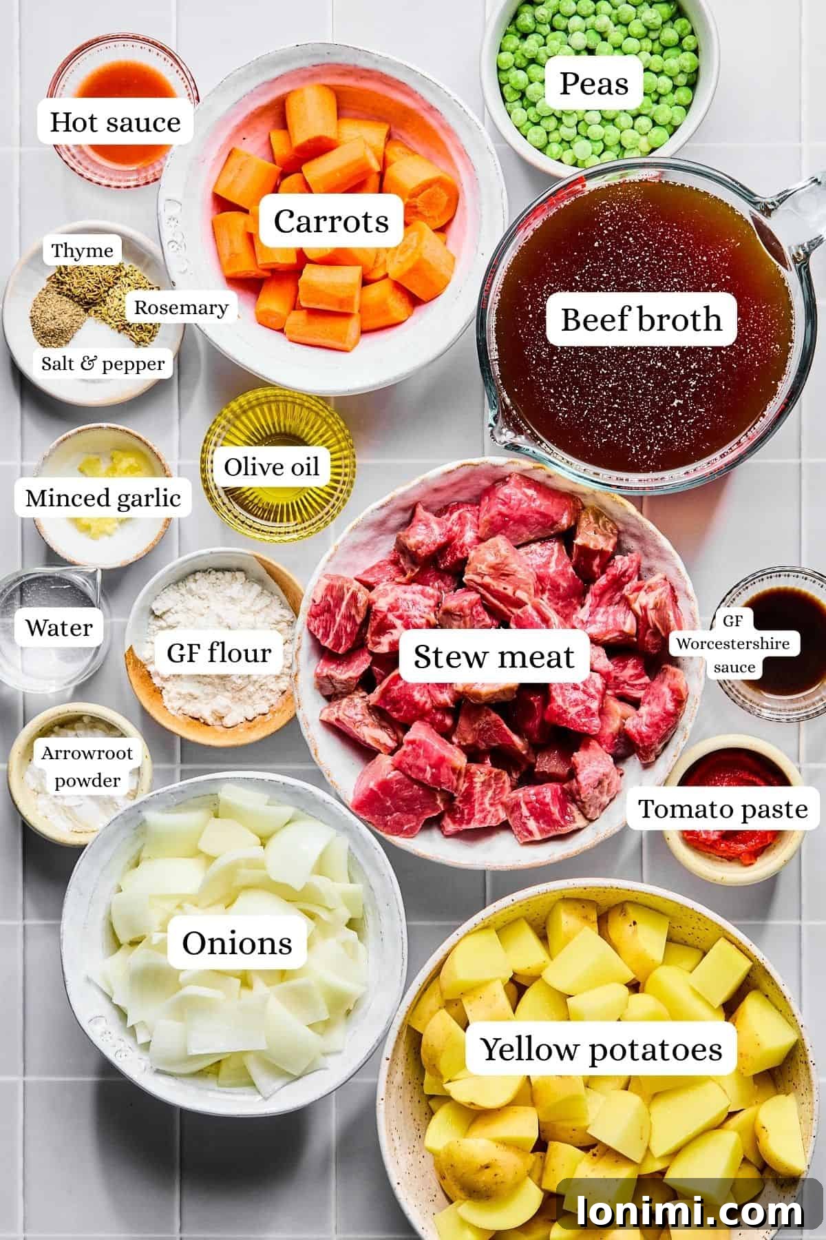 Slow cooker beef stew ingredients laid out in nesting bowls on a white countertop, including chopped beef, carrots, potatoes, and spices.